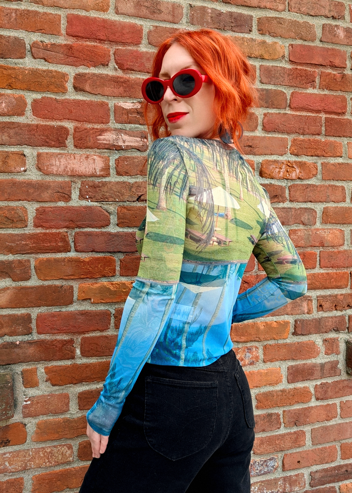 Person wearing a colorful long-sleeve mesh shirt with scenic design and red sunglasses against a brick wall.