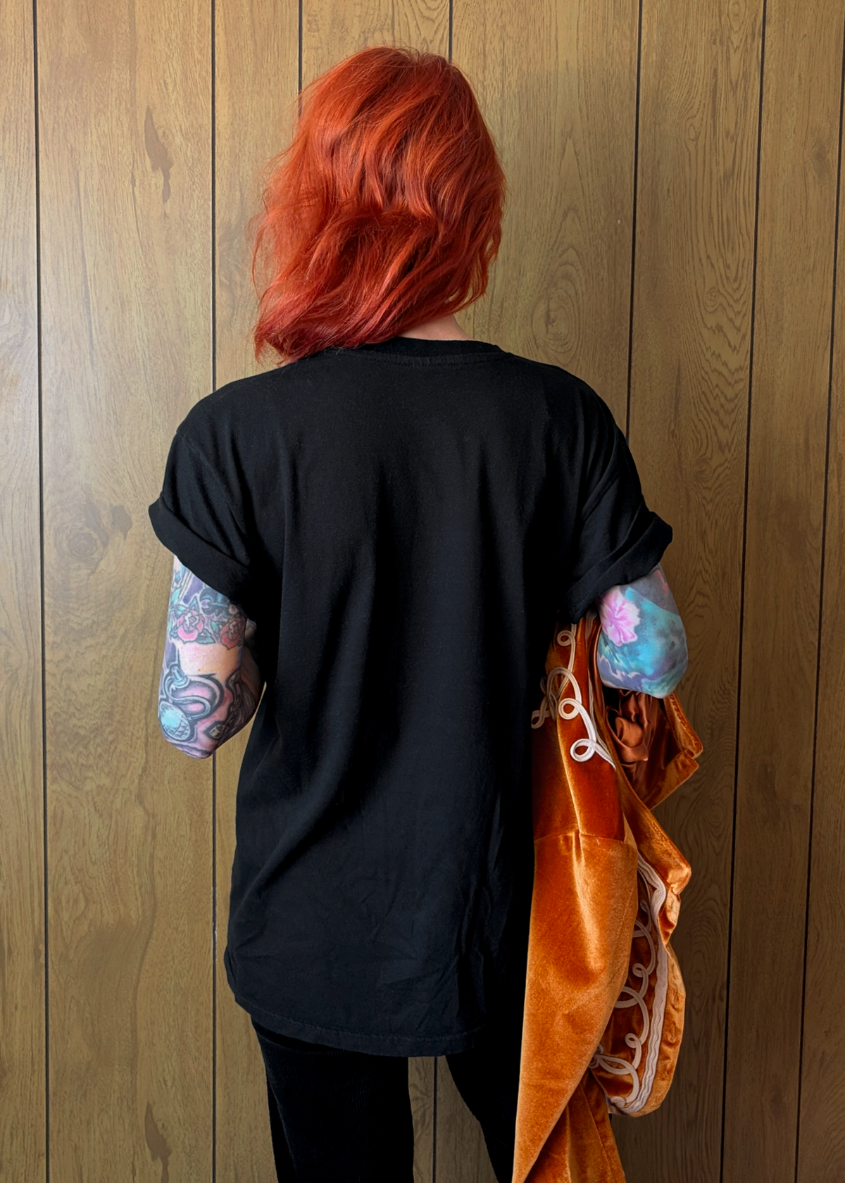Person with red hair wearing a black shirt and holding an orange jacket against a wooden wall.