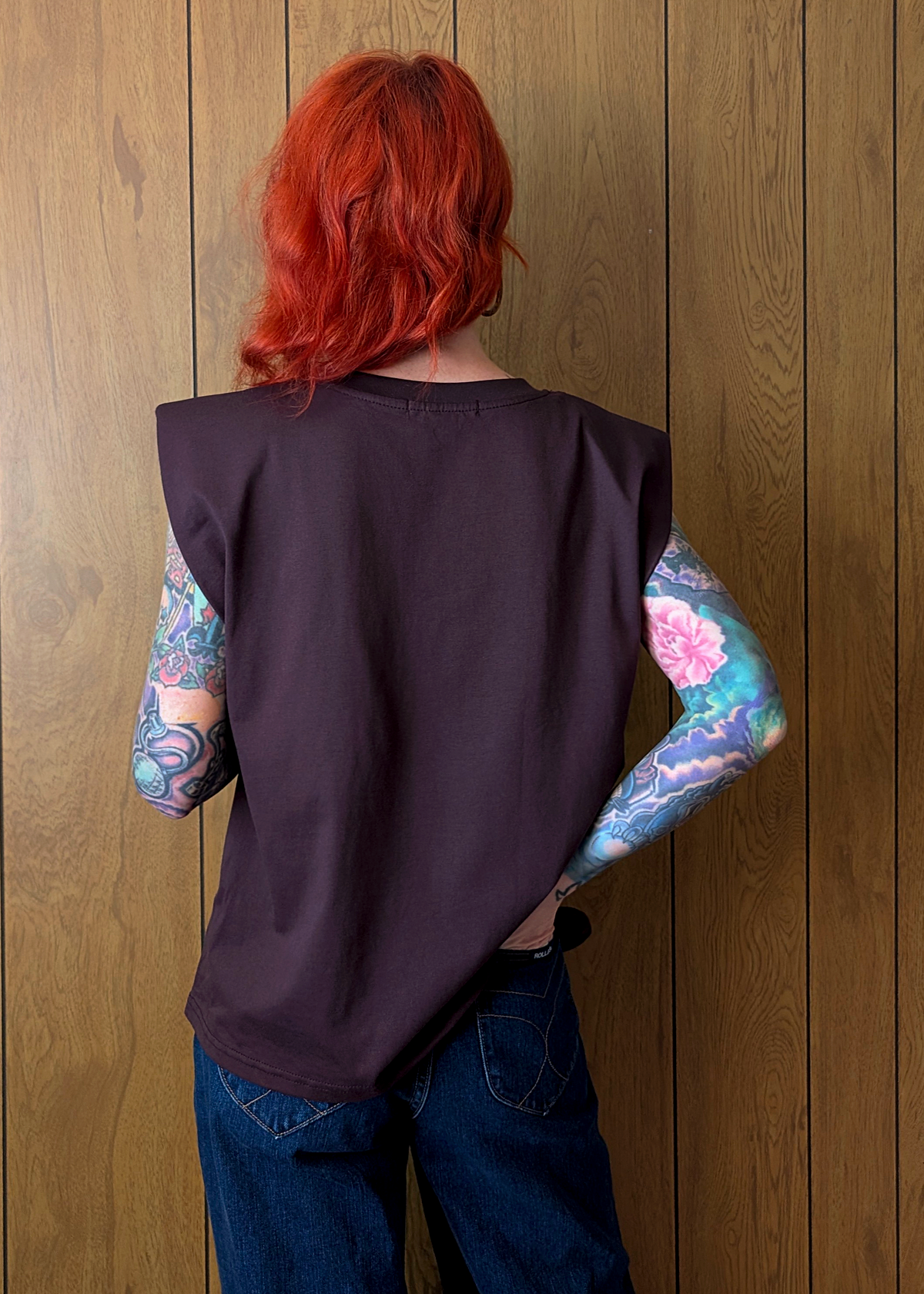 Person with red hair and colorful tattoos on arms wearing a dark plum sleeveless top against a wooden wall.