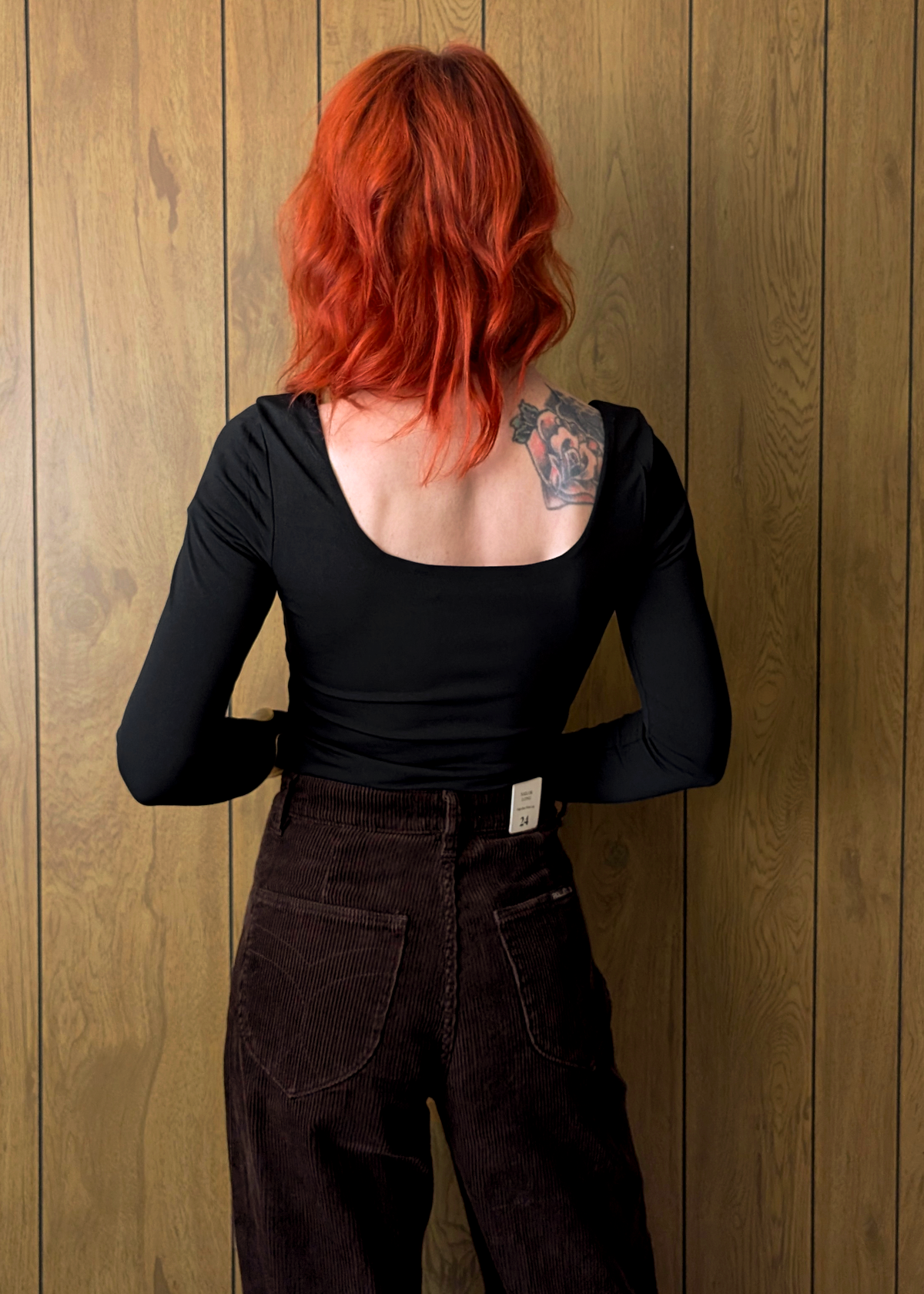 Person with red hair and a tattoo on their back, wearing a black top and dark brown cord pants, standing against a wooden wall.