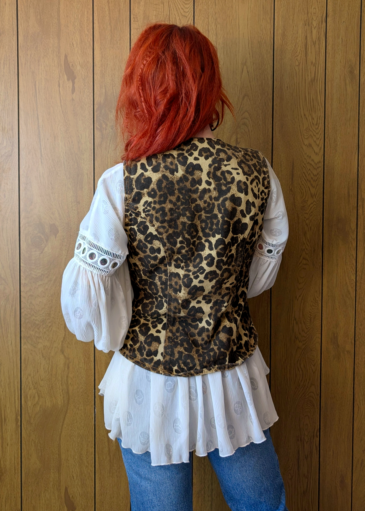 Person wearing a leopard print vest over a white blouse with blue jeans against a wooden panel background