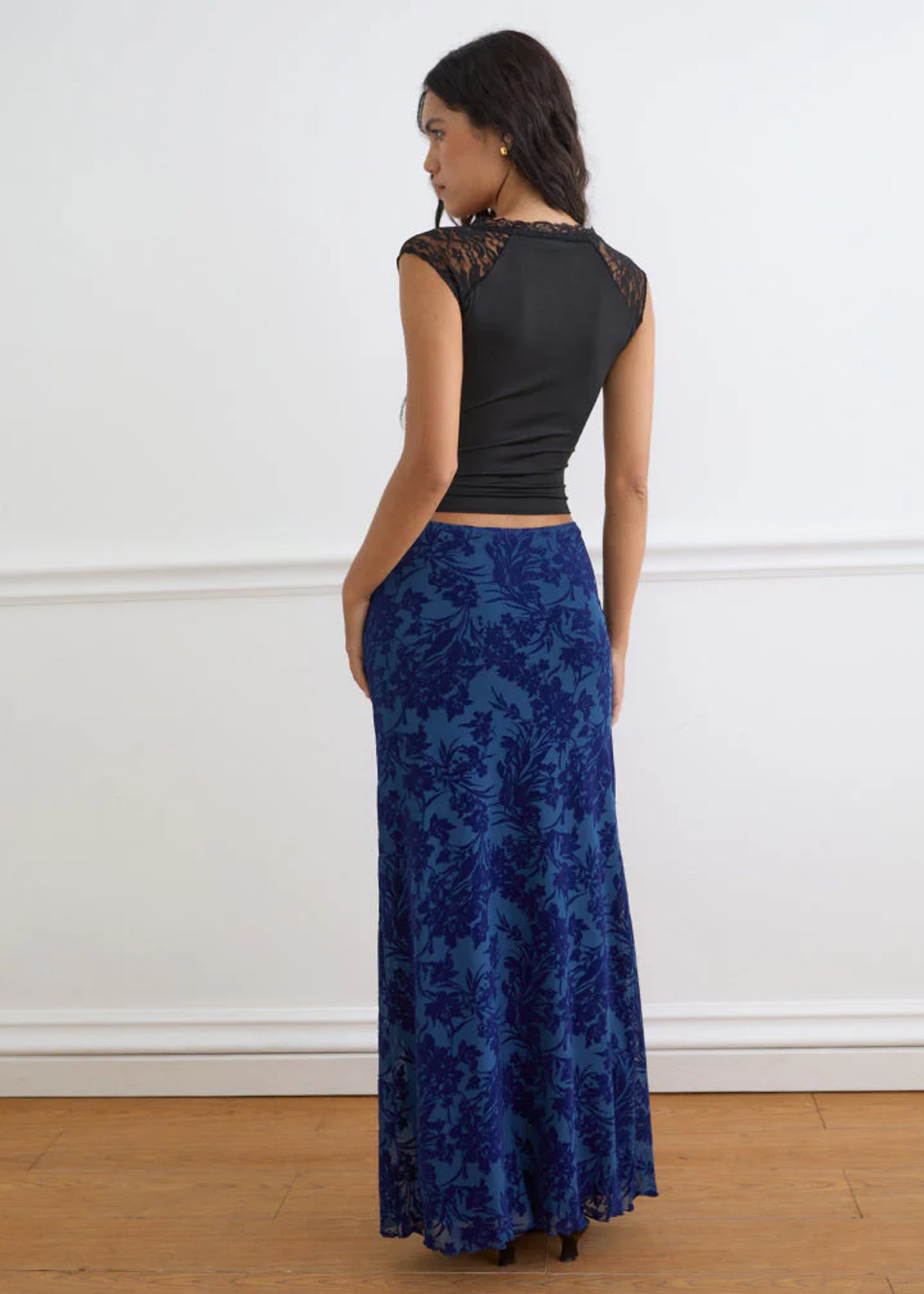 Woman wearing a black top and blue floral skirt against a white wall.