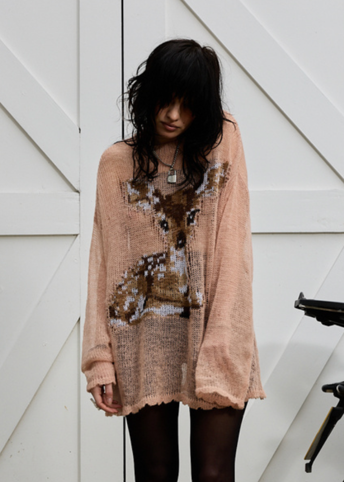 Woman wearing a dusty pink open knit lightweight sweater with deer design with black tights, posed against a white wooden background