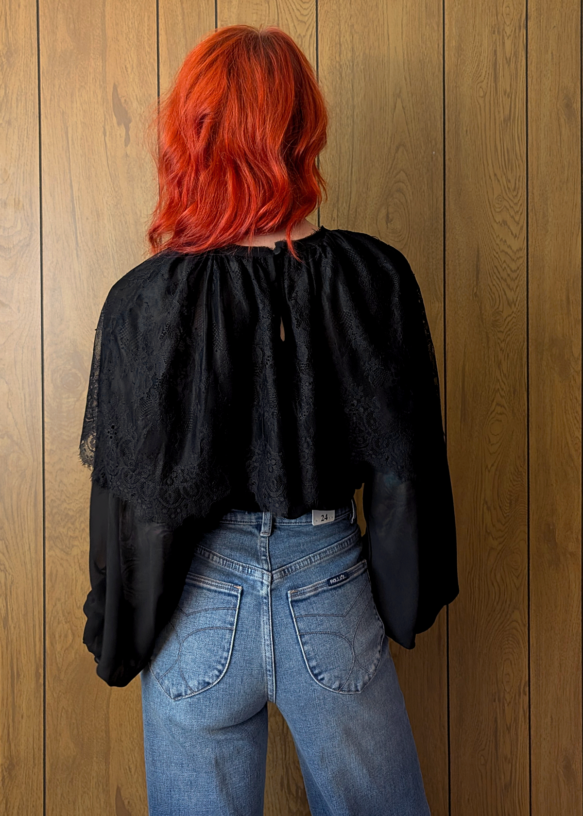 Person with red hair wearing a black top and blue jeans against a wooden wall.