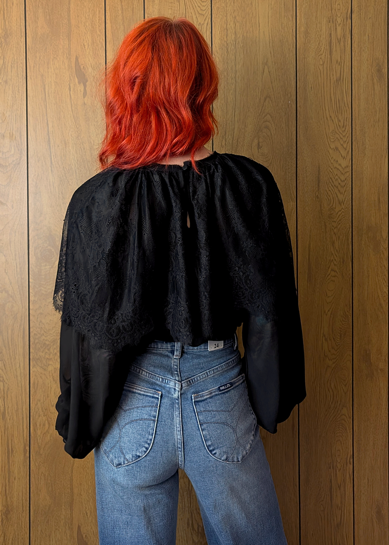 Person with red hair wearing a black top and blue jeans against a wooden wall.