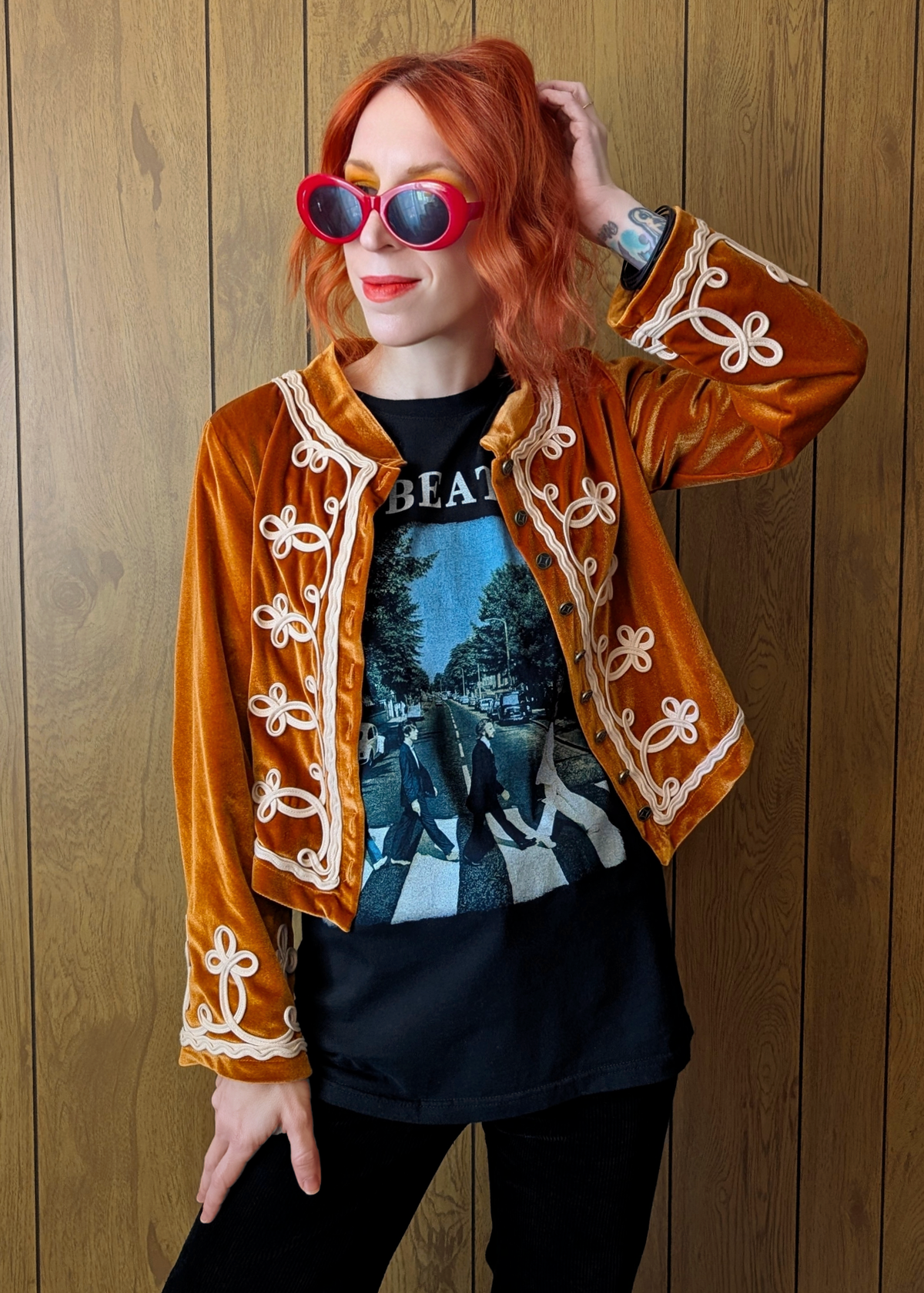 Person wearing an orange velvet jacket with white embroidery, black Beatles t-shirt with graphic design, and red sunglasses against a wooden background.