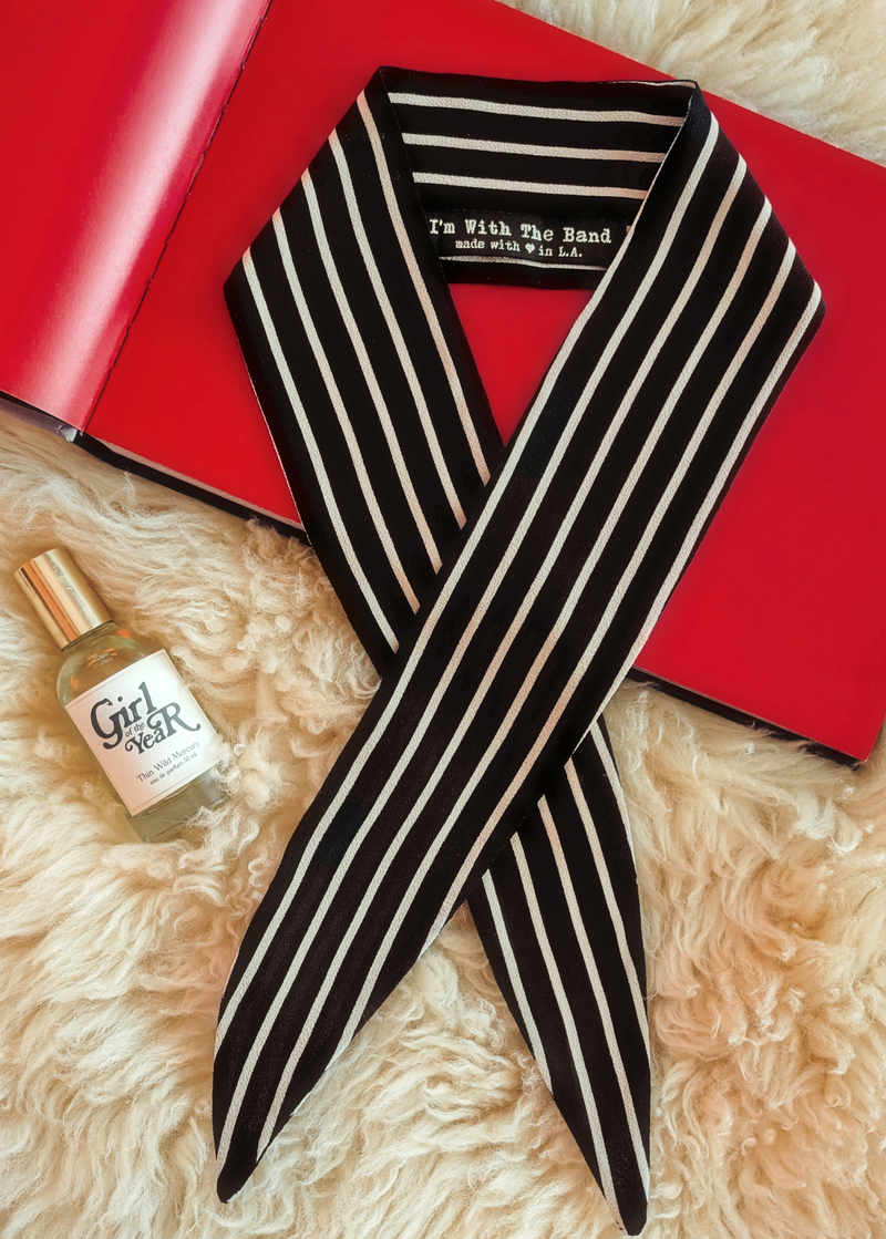 Lifestyle photo of a black and beige stripe scarf laid flat on a cream faux fur rug with a red book and bottle of perfume