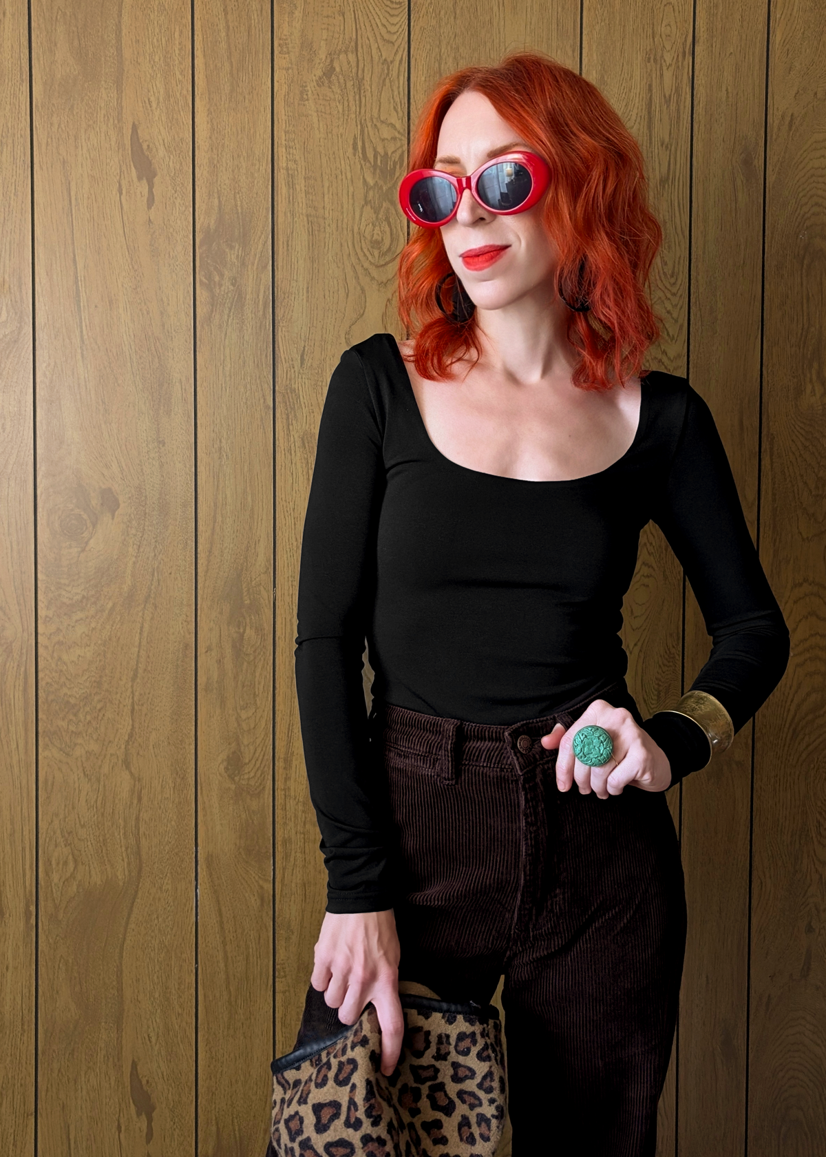 Woman with red hair wearing sunglasses and a black top against a wooden wall.