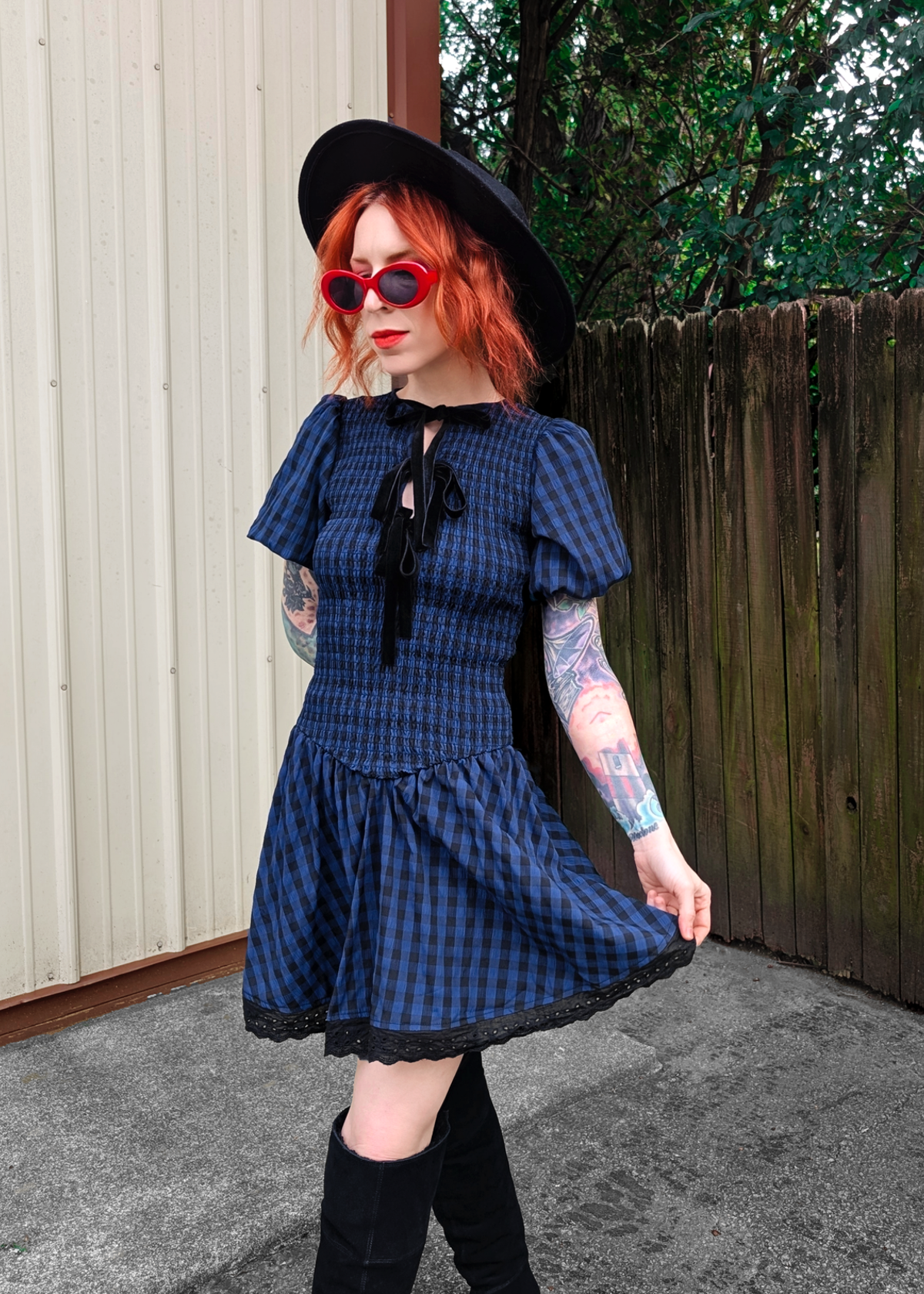Person wearing a blue and black checkered dress with puff sleeves, black hat, and red sunglasses outdoors.
