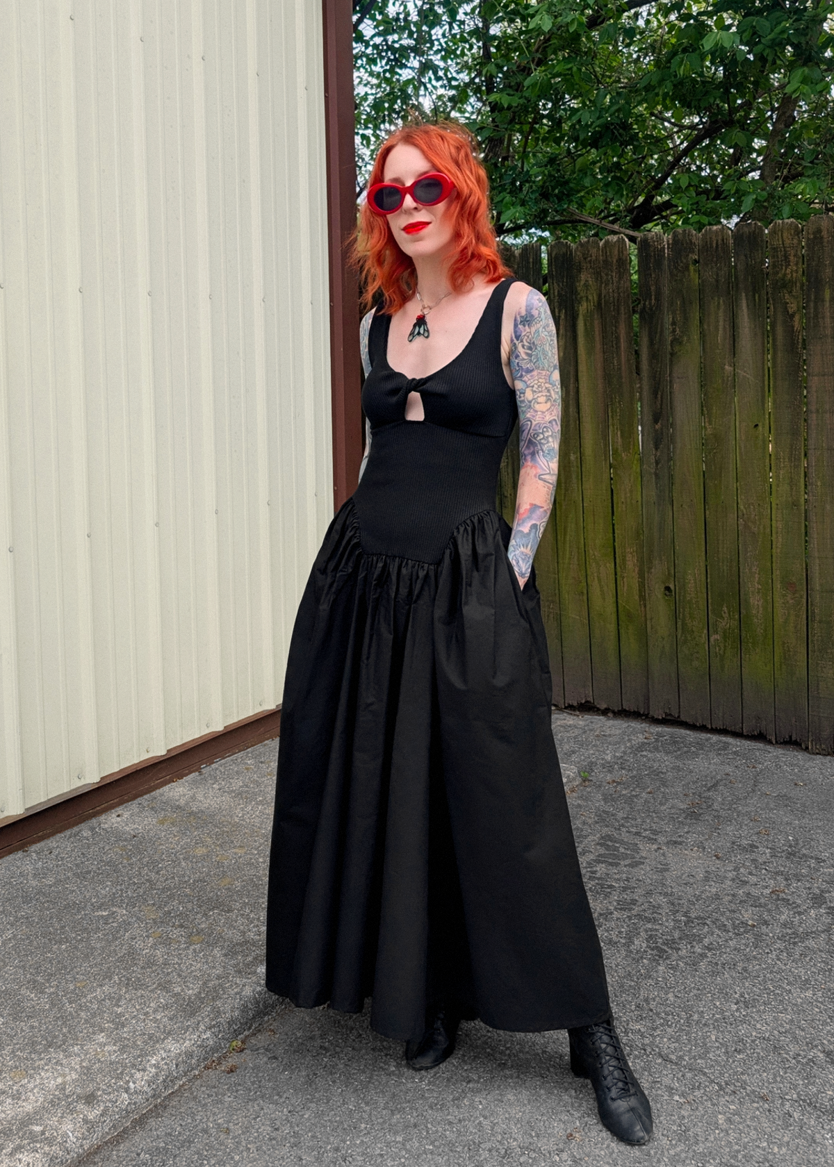 Red headed lady wearing a black sleeveless cotton maxi dress outside against a wood fence