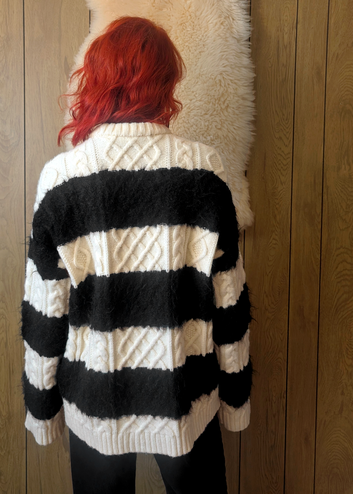 Person wearing a black and cream striped cable knit sweater against a wooden wall.