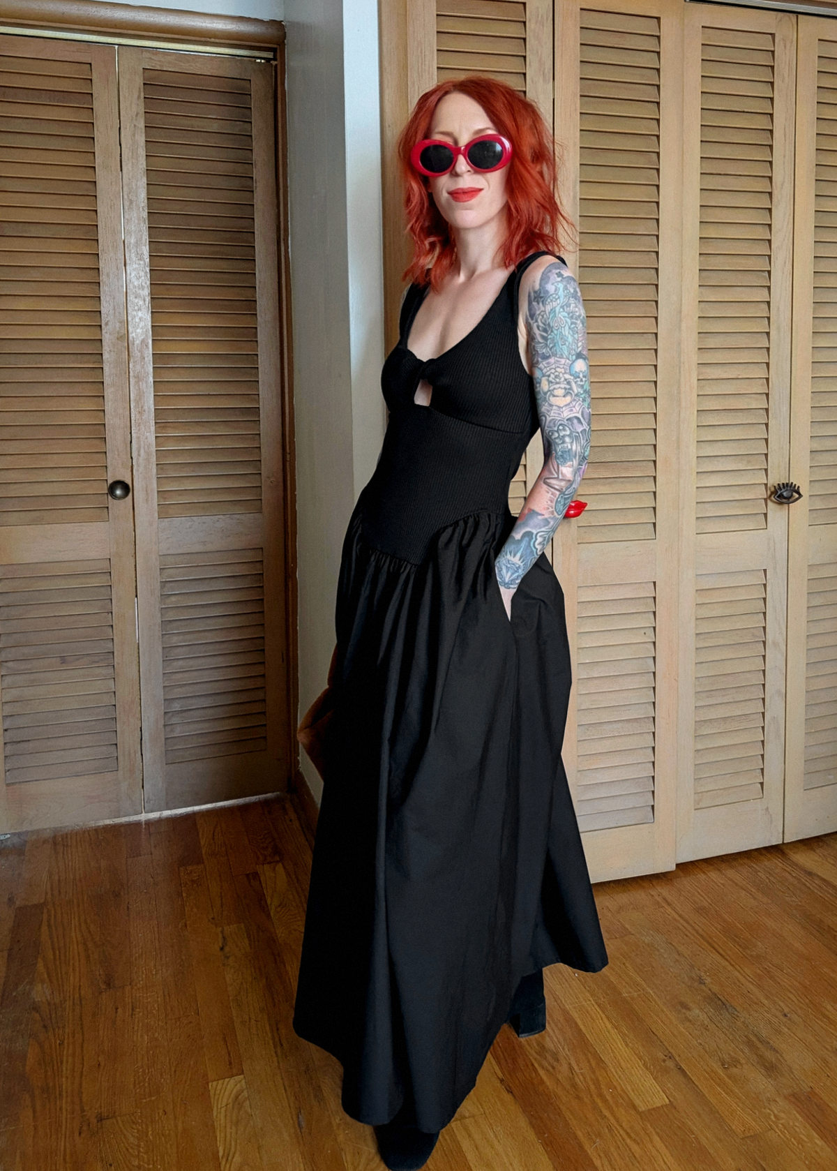 Person with red hair and tattoos wearing a black midi maxi dress standing in a room with wooden floors and shutters.