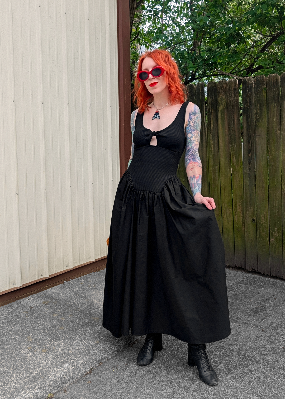 Person wearing a black dress with red hair and sunglasses standing outdoors.