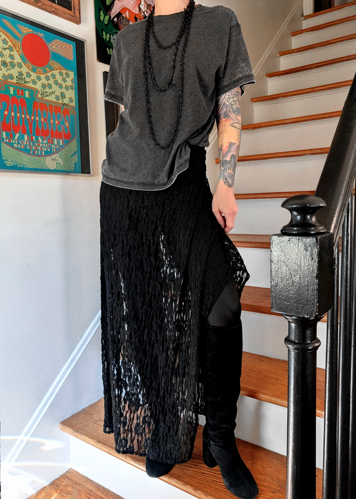 A person wearing a burnout black oversized t-shirt, a long black lace maxi skirt over black leggings and black knee-high boots, standing on wooden stairs in a home interior.