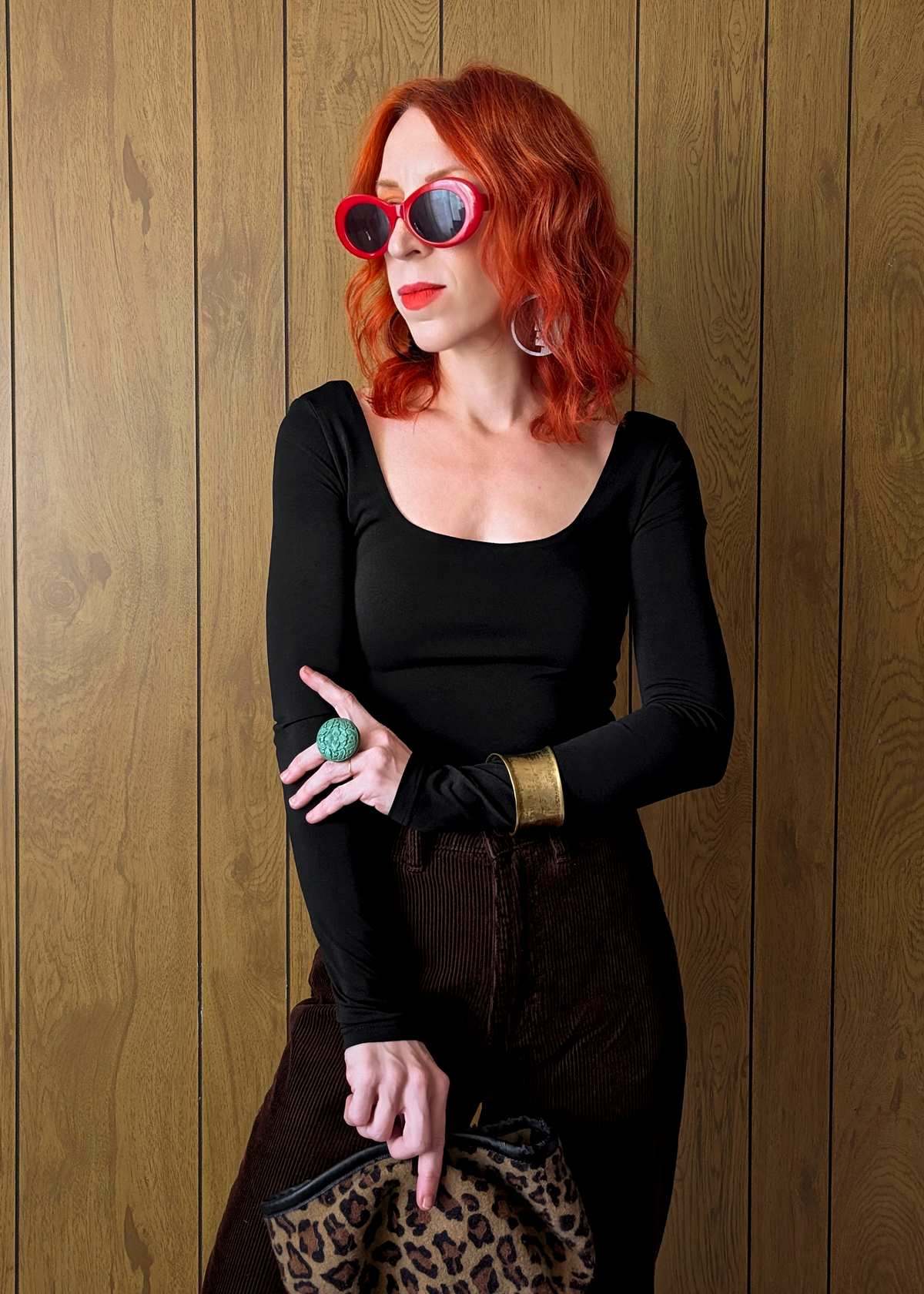 Woman with red hair wearing sunglasses and a black top against a wooden wall.