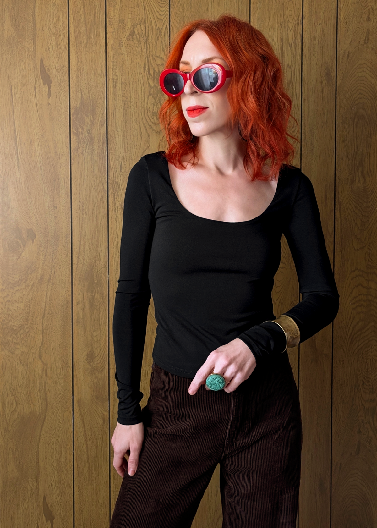 Person with red hair wearing black top and brown pants against a wooden wall