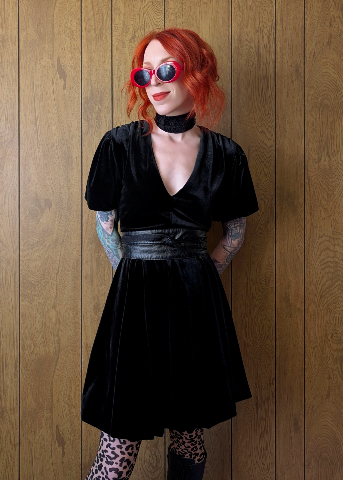 Person wearing a black velvet mini dress with red hair and sunglasses against a wooden wall.