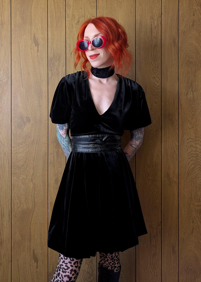 Person wearing a black velvet mini dress with red hair and sunglasses against a wooden wall.