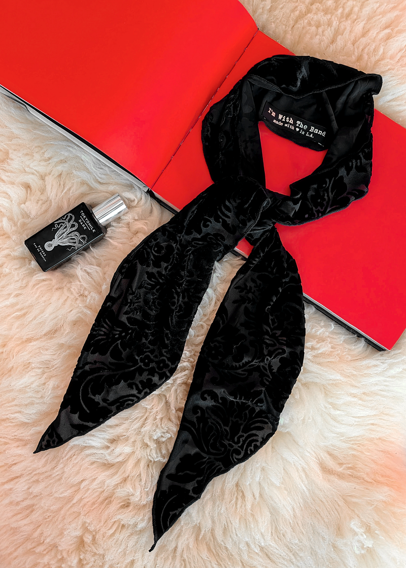 Lifestyle photo of a black burnout velvet scarf laid flat on a cream faux fur rug with a red book and bottle of perfume