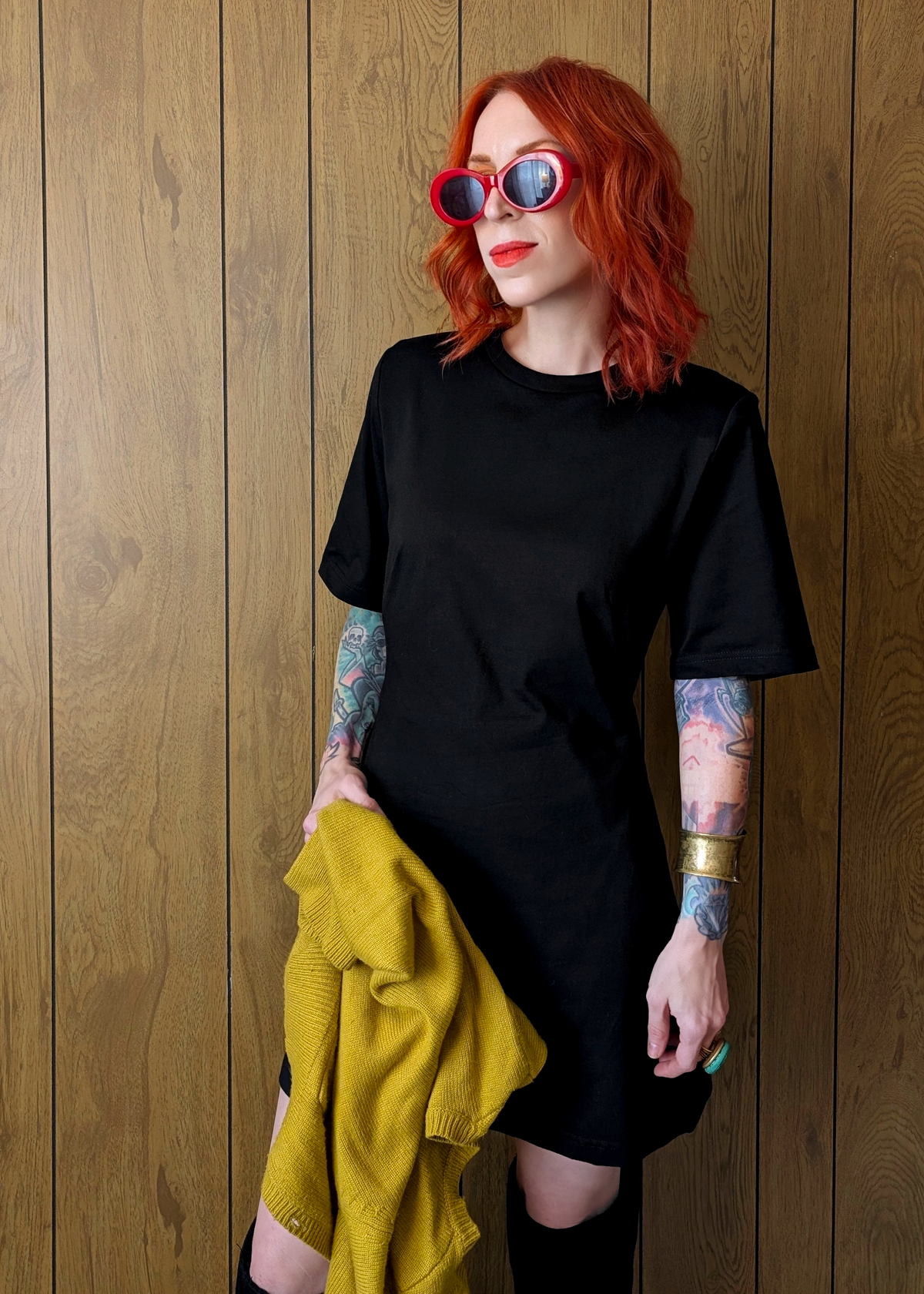 Person with red hair wearing a black dress and sunglasses, holding a yellow jacket against a wooden wall.