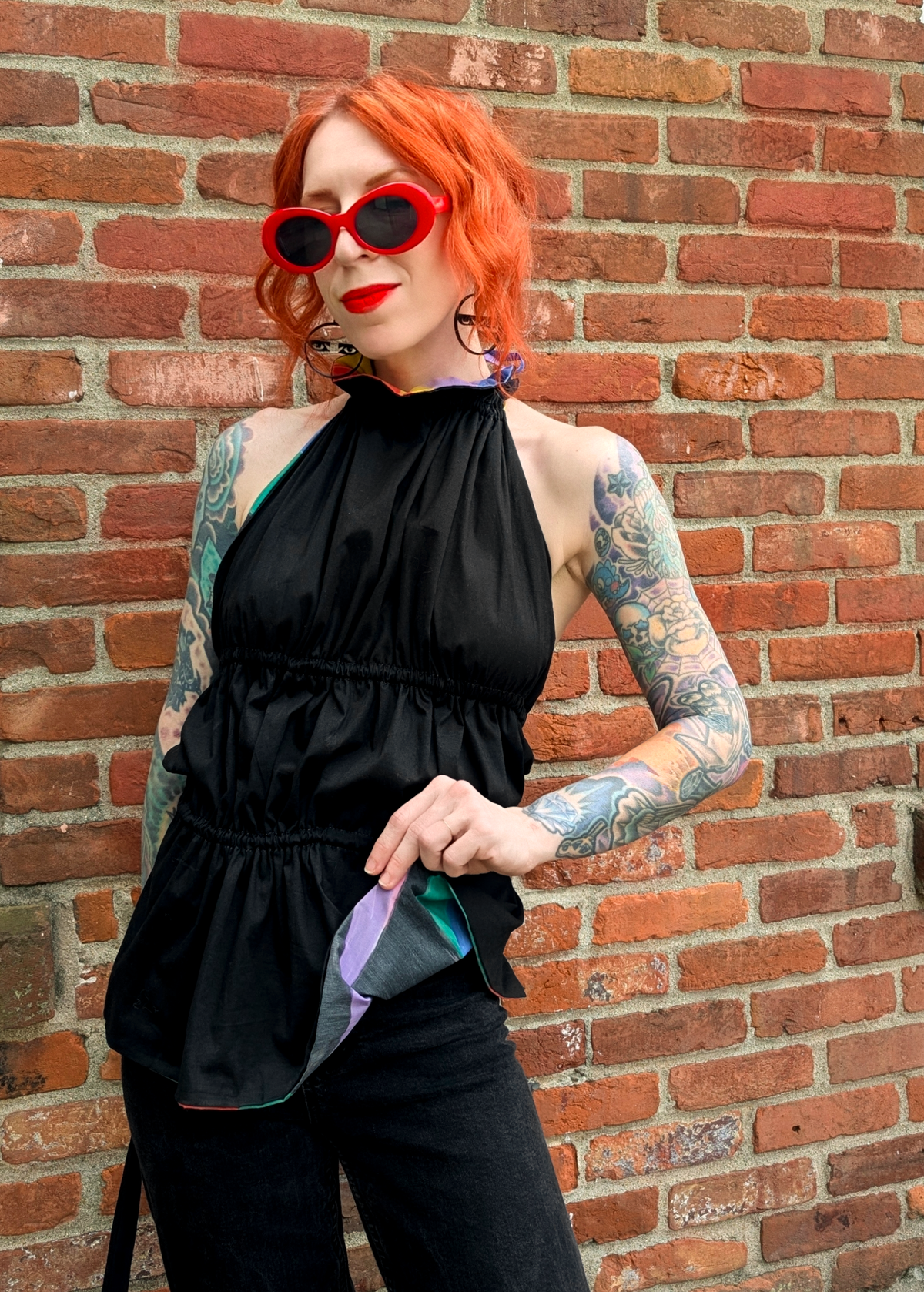 Person with tattoos and red hair wearing a black halter top with black jeans against a brick wall.
