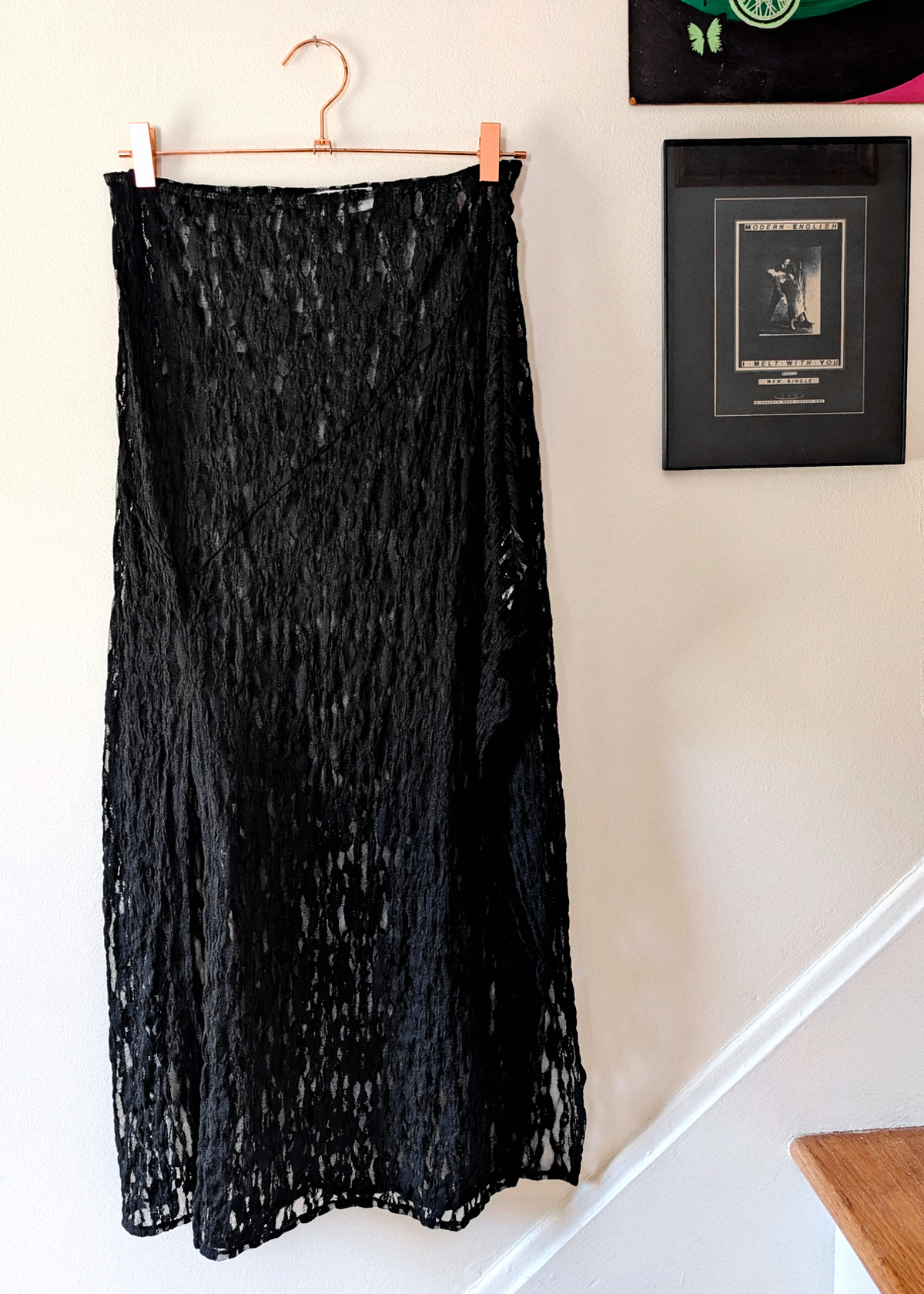 A sheer black lace midi skirt hanging from a copper metal hanger with clips on a light-colored wall next to framed art.