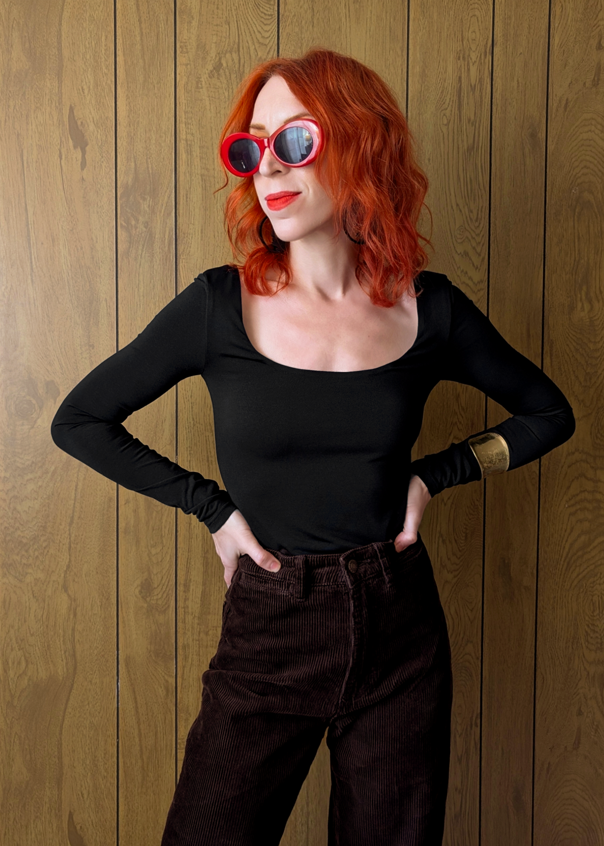 Person wearing a black long-sleeve top and brown corduroy pants with red sunglasses against a wooden background
