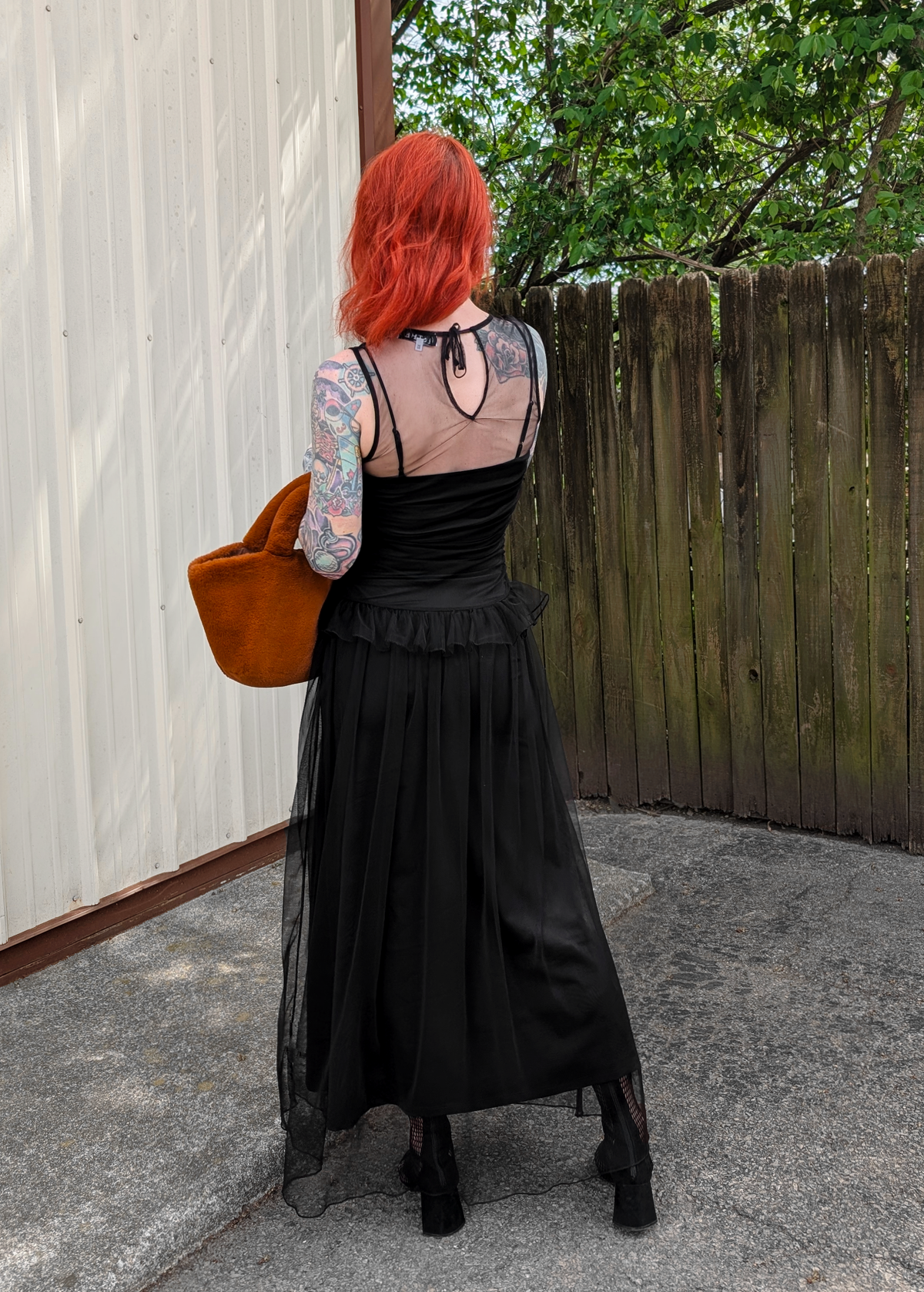 Person with red hair wearing a black dress and carrying a brown faux fur bag, standing in front of a wooden fence.