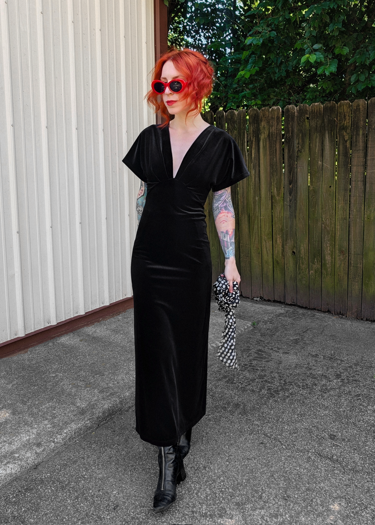 woman with red hair wearing a black velvet short sleeve midiaxi dress with black boots and wearing red sunglasses outside