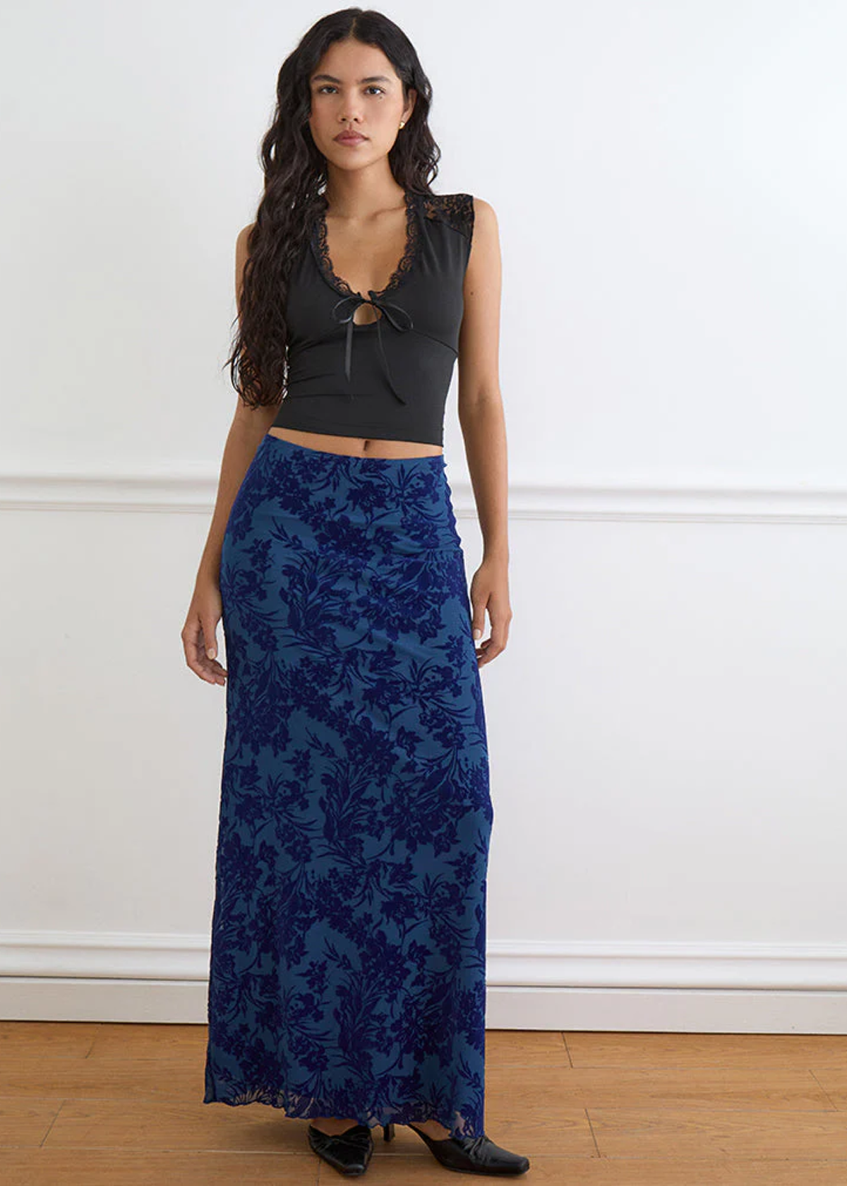Woman wearing a black top and blue floral skirt against a white wall.
