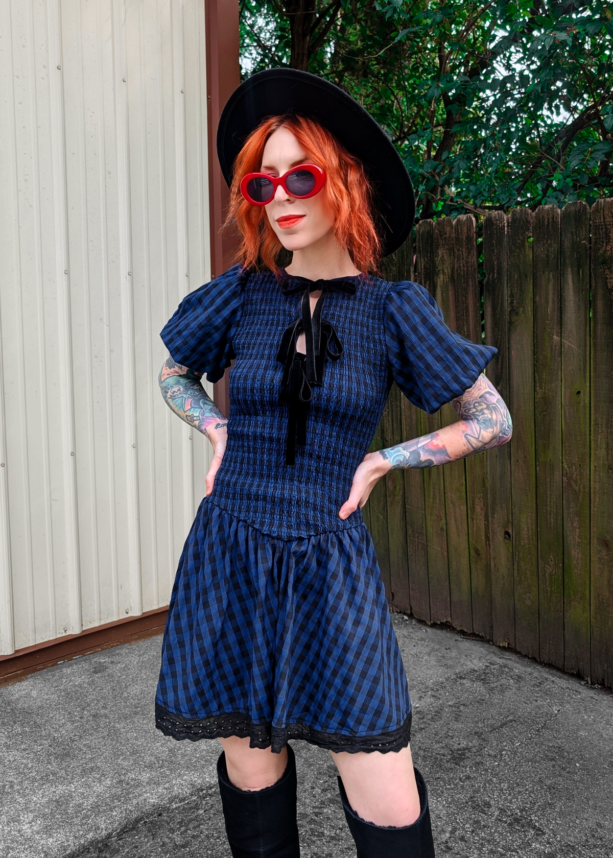 Person wearing a blue and black  checkered dress with puffed sleeves, black hat, and sunglasses outdoors.