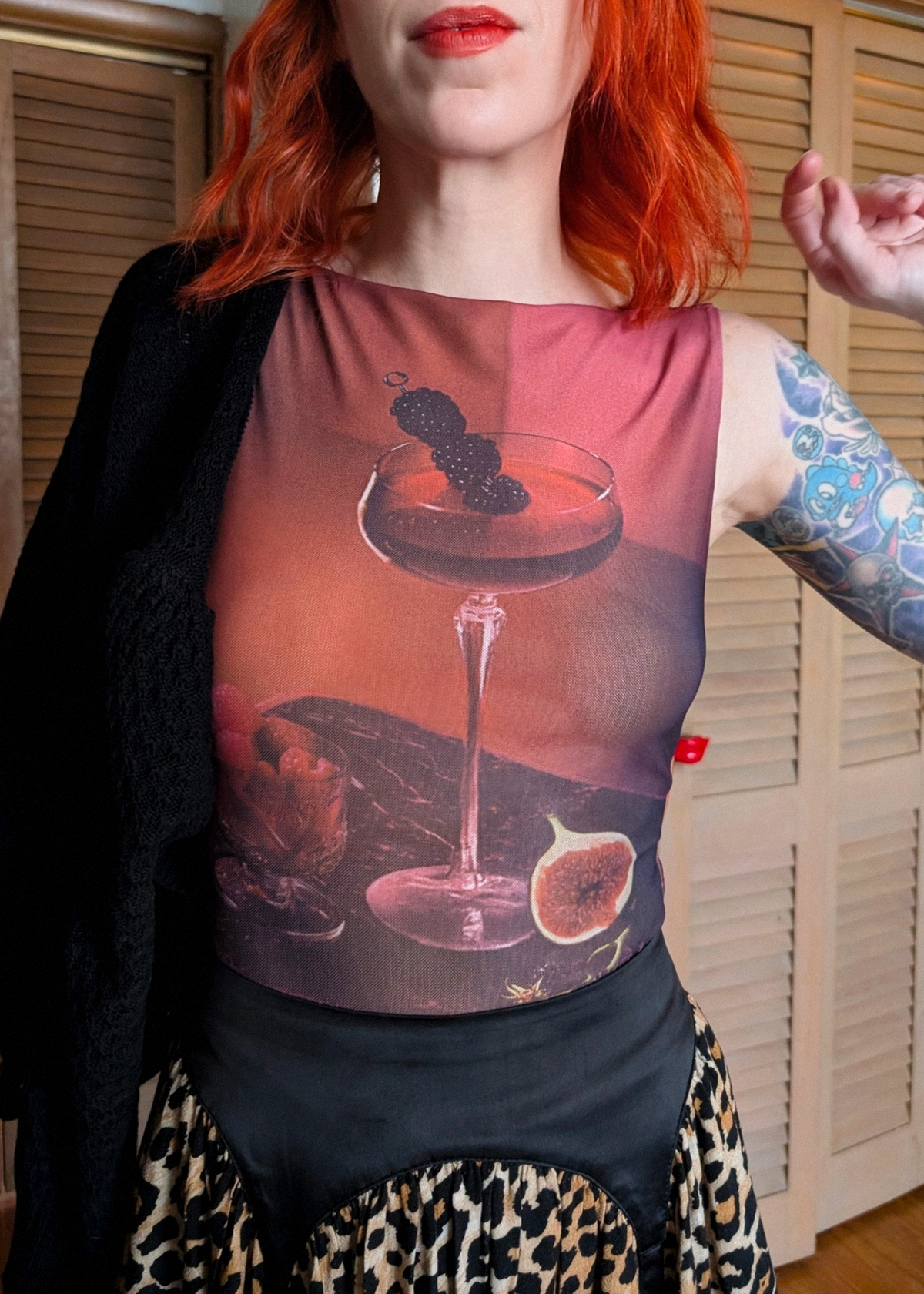 A person wearing a sleeveless top with a large printed graphic of a cocktail glass, fig, and dark fruit on a warm pink-brown background, paired with a black cardigan and leopard-print skirt, standing indoors in front of wooden shutters.