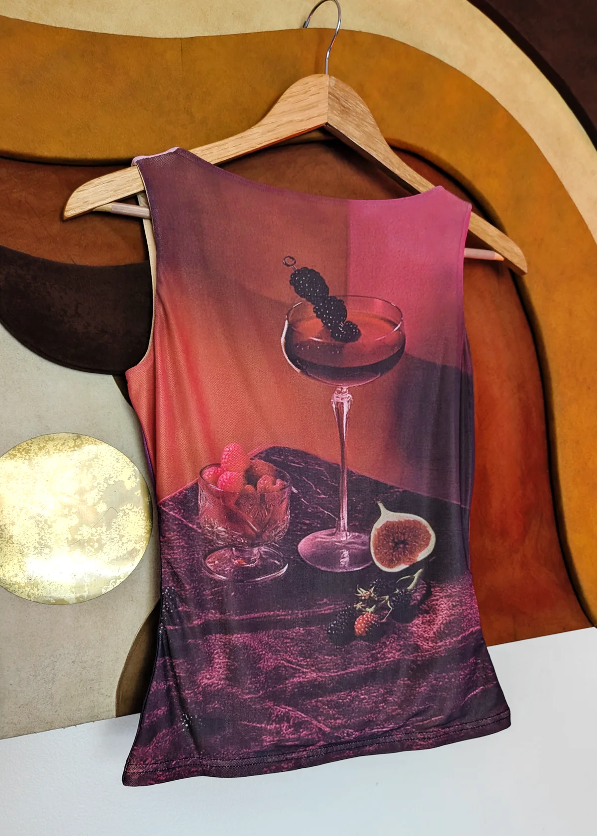 A sleeveless scoop-neck mesh tank top hanging on a wooden hanger, featuring an all-over photographic print of a cocktail glass with a blackberry, strawberries, and a fig 