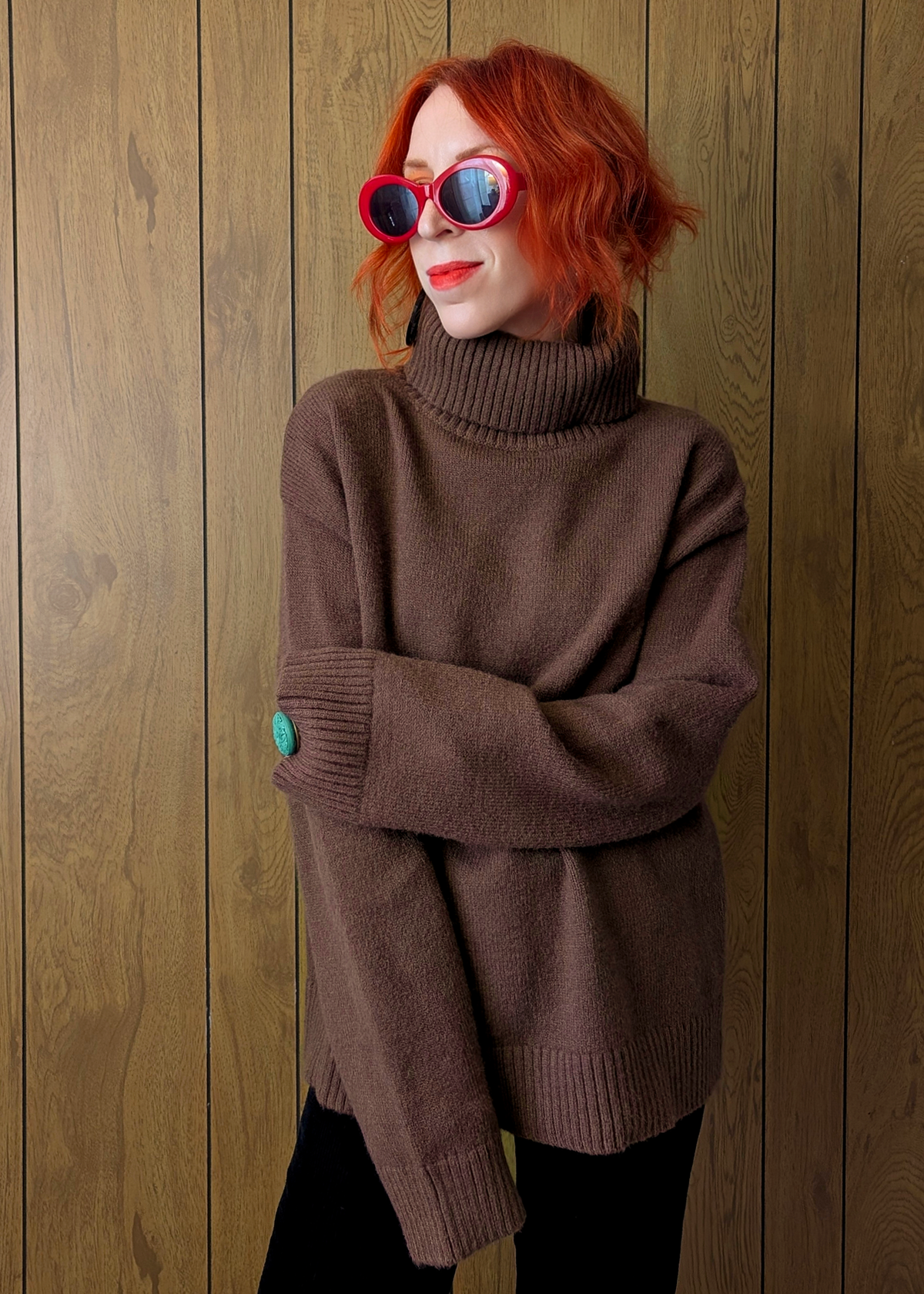 Person wearing a brown sweater and red sunglasses against a wooden wall