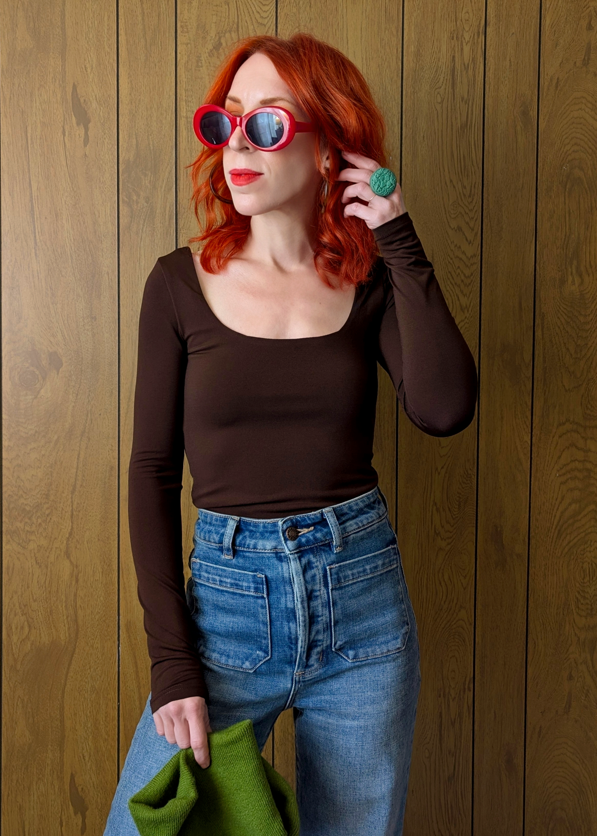 Woman with red hair wearing sunglasses and a brown top against a wooden wall