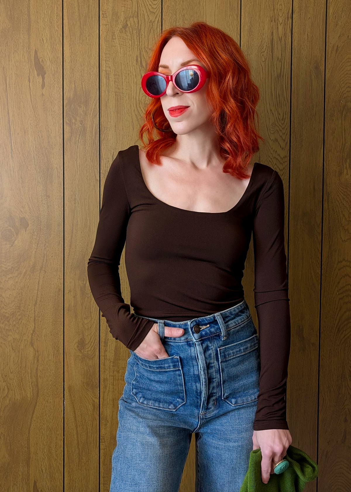 Woman with red hair wearing sunglasses and a brown top against a wooden wall