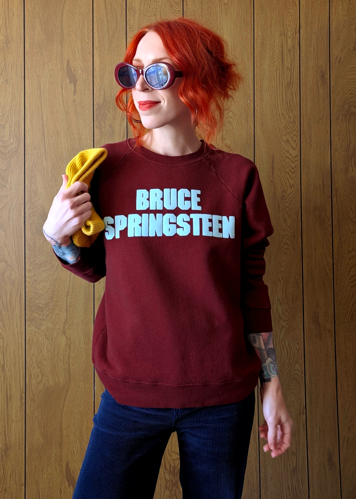 Person wearing a maroon sweatshirt with 'BRUCE SPRINGSTEEN' text, holding a beanie against a wooden background.