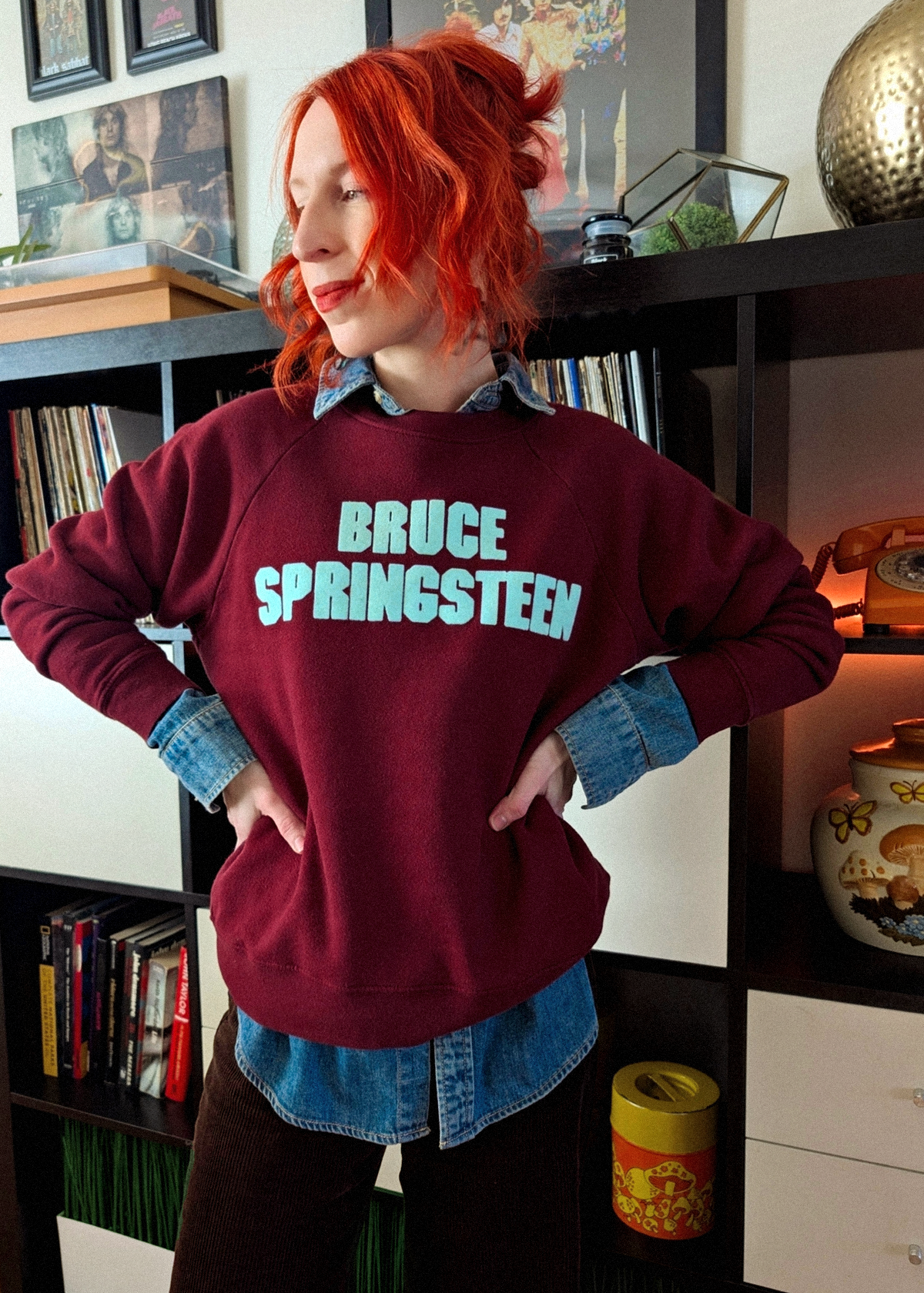 Person wearing a maroon sweatshirt with 'Bruce Springsteen' text in a room with bookshelves and decorative items.