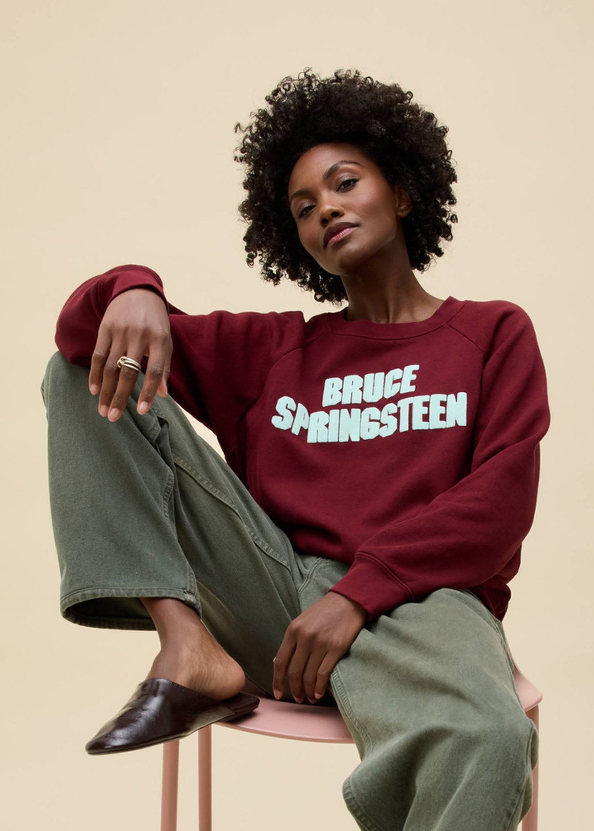 Person wearing a maroon sweatshirt with 'Bruce Springsteen' text, sitting on a chair against a beige background
