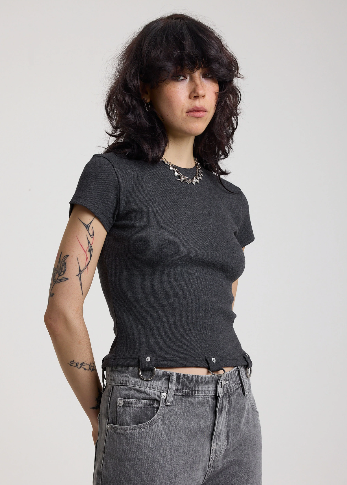 Woman wearing a ribbed charcoal grey shirt with D-ring details at hem, styled with light grey jeans, posed against a white backdrop