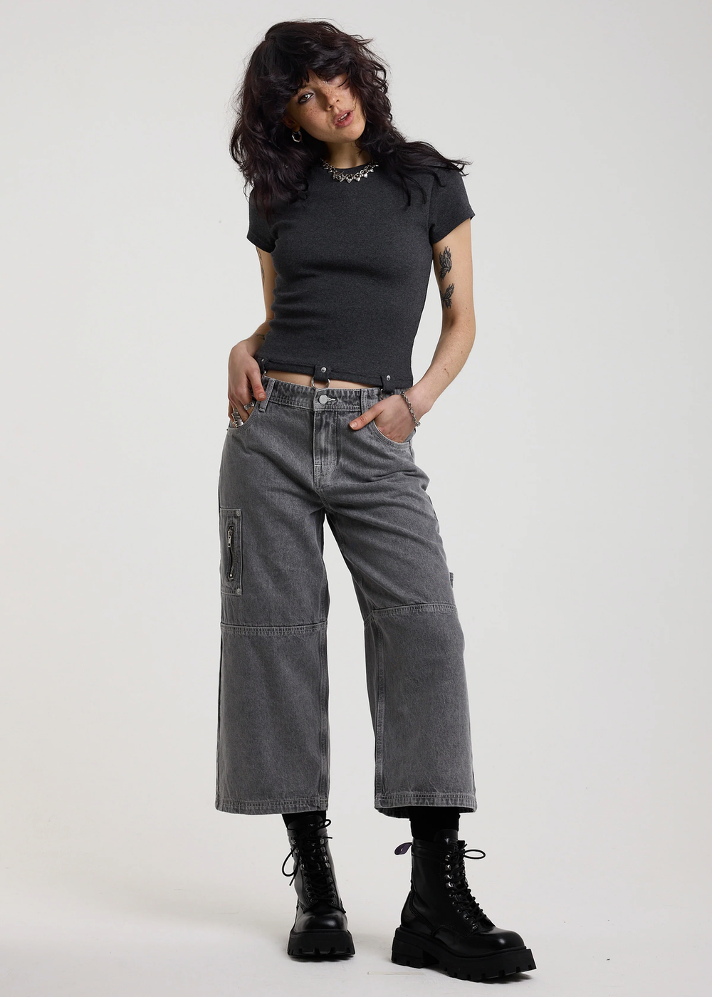 Woman wearing a ribbed charcoal grey shirt with D-ring details at hem, styled with light grey jeans, posed against a white backdrop