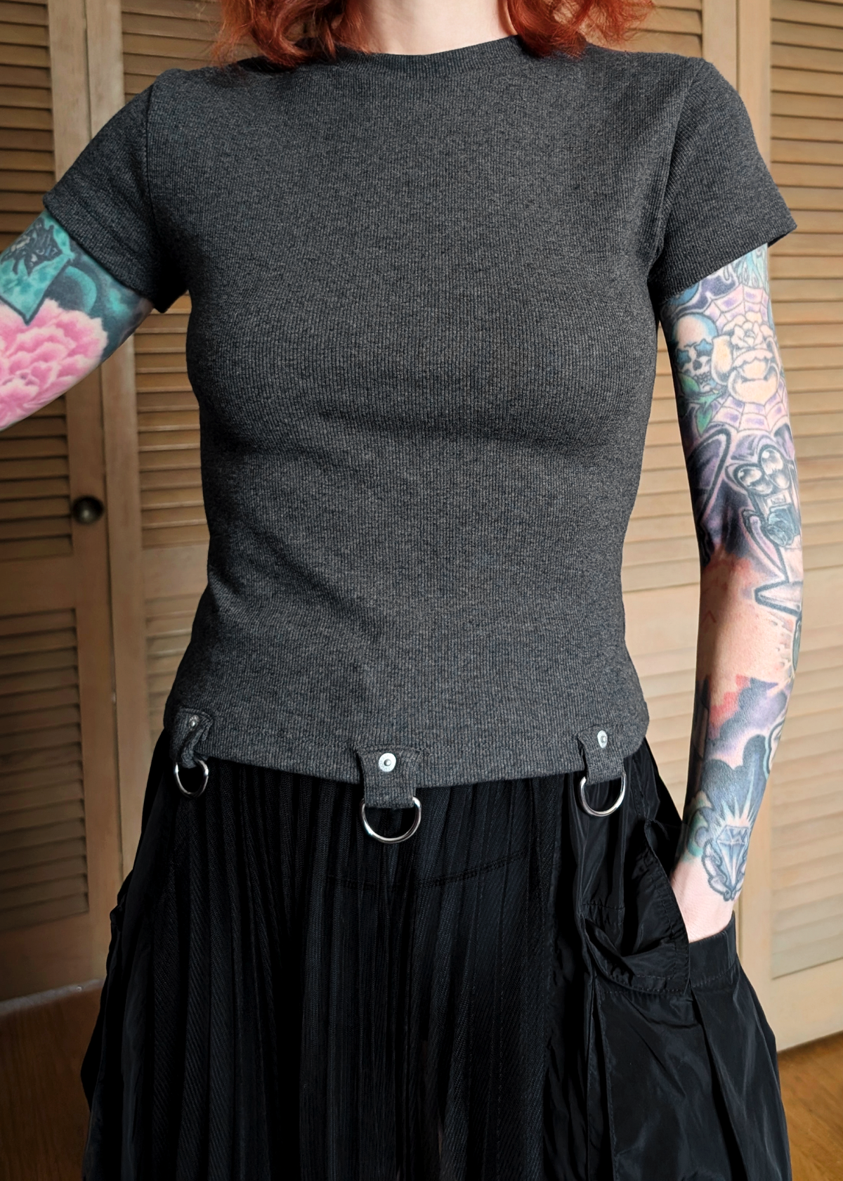 Person wearing a ribbed charcoal grey t-shirt with D-ring details at hem, and black skirt standing indoors.