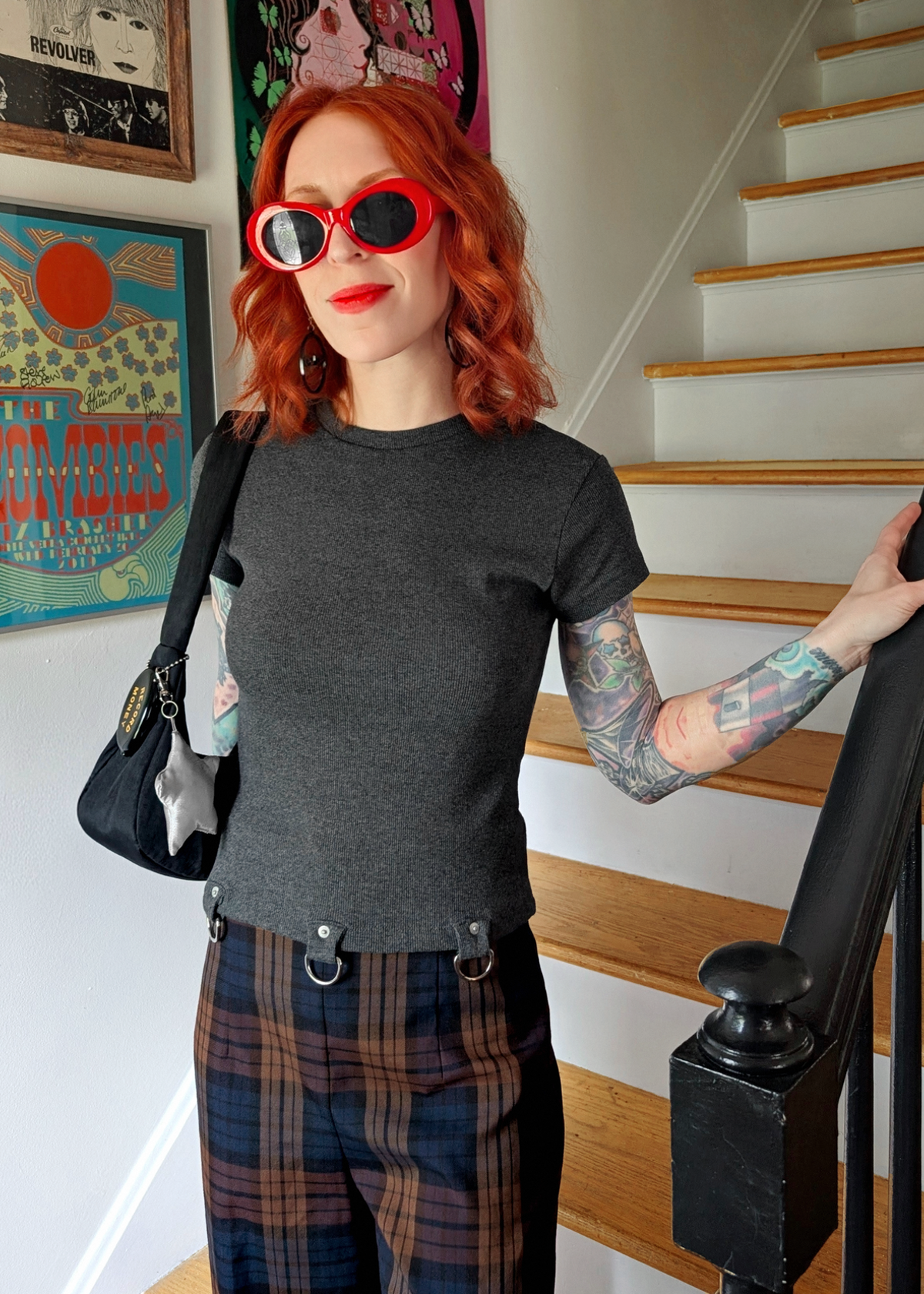 Person with red hair and tattoos wearing a ribbed charcoal grey shirt with D ring details at hem and plaid pants, standing in a room with a staircase.