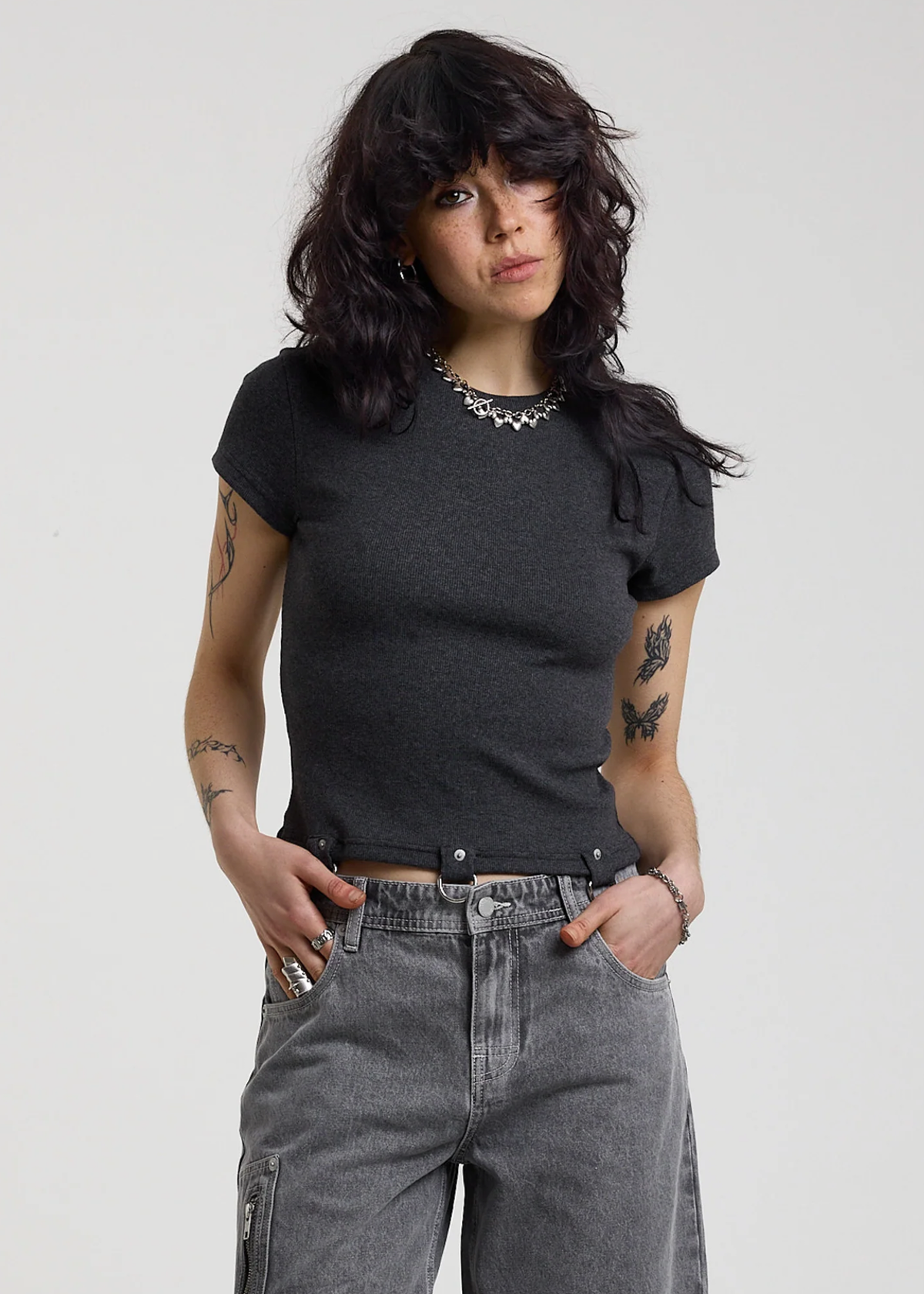 Woman wearing a ribbed charcoal grey shirt with D-ring details at hem, styled with light grey jeans, posed against a white backdrop