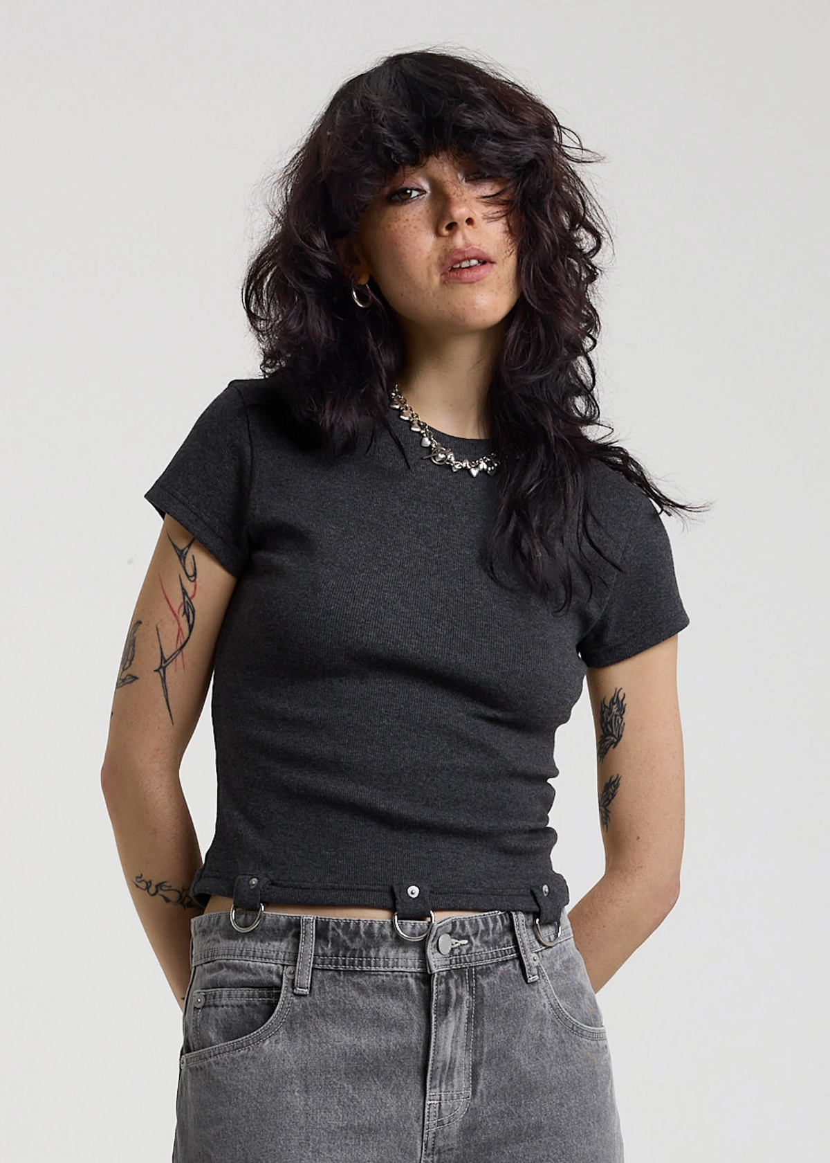 Woman wearing a ribbed charcoal grey shirt with D-ring details at hem, styled with light grey jeans, posed against a white backdrop