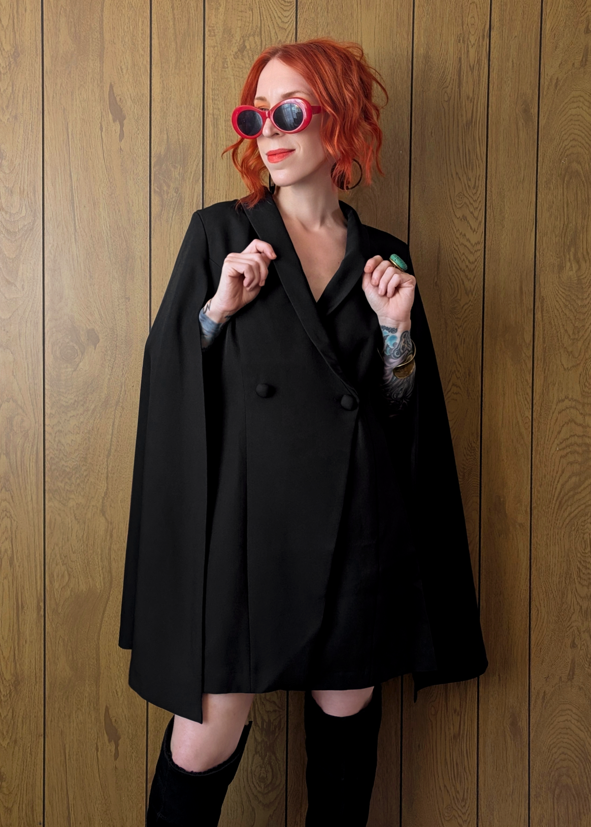 Person wearing a black cape blazer dress and sunglasses against a wooden wall