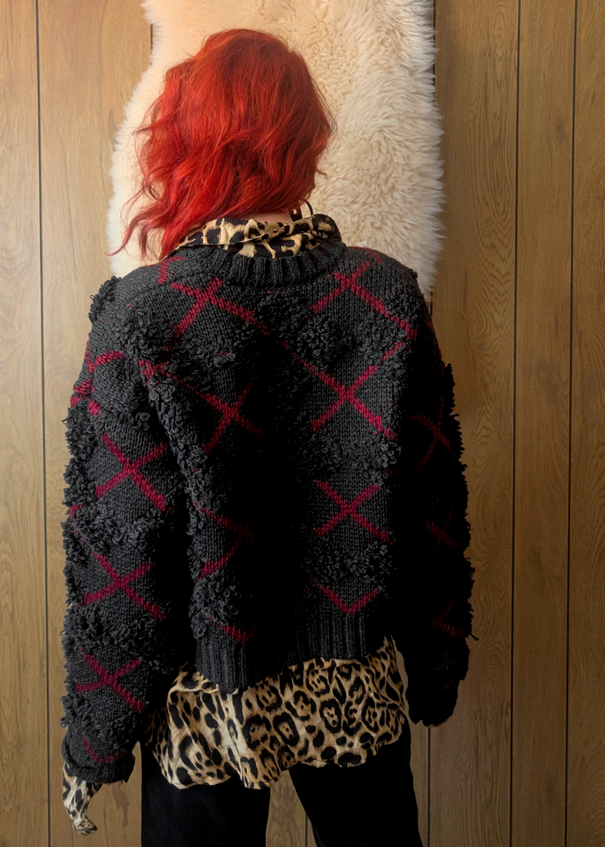 Person wearing a charcoal and red argyle "x" patterned cardigan over a leopard print shirt against a wooden wall.