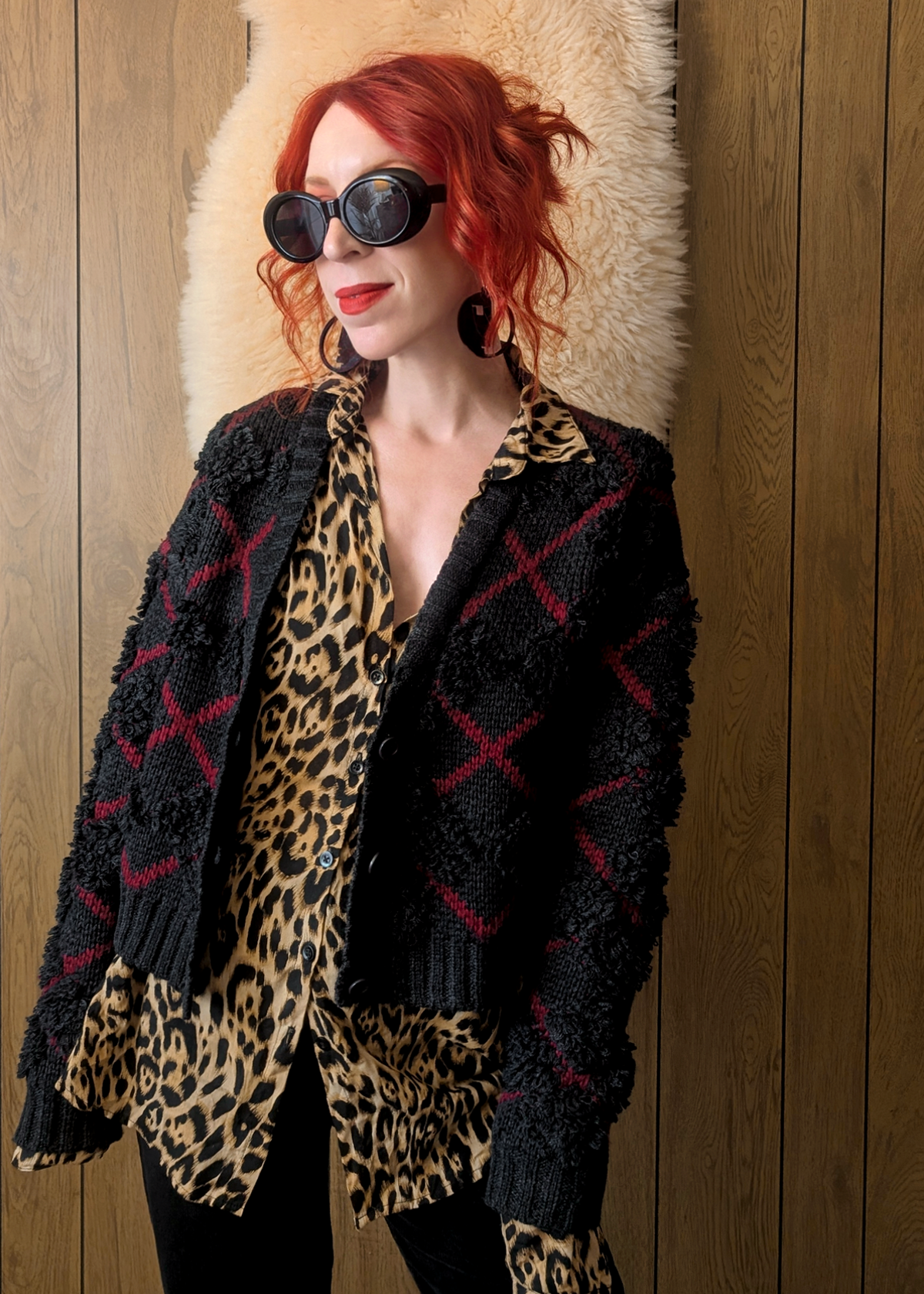 Person wearing a charcoal and red argyle "x" patterned cardigan over a leopard print shirt against a wooden wall.