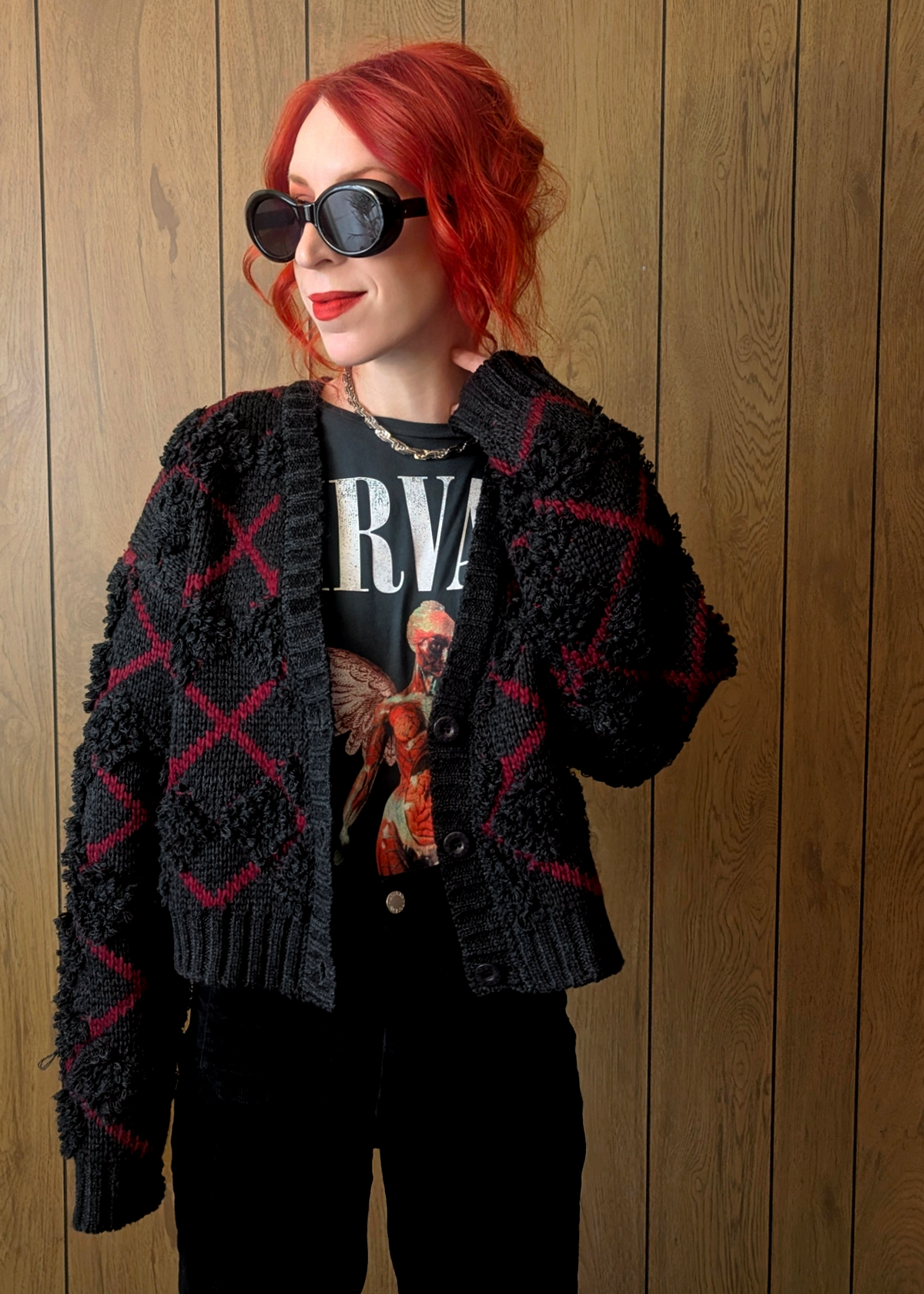 Person wearing a charcoal textured cardigan with red "x" argyle pattern over a graphic t-shirt against a wooden panel background
