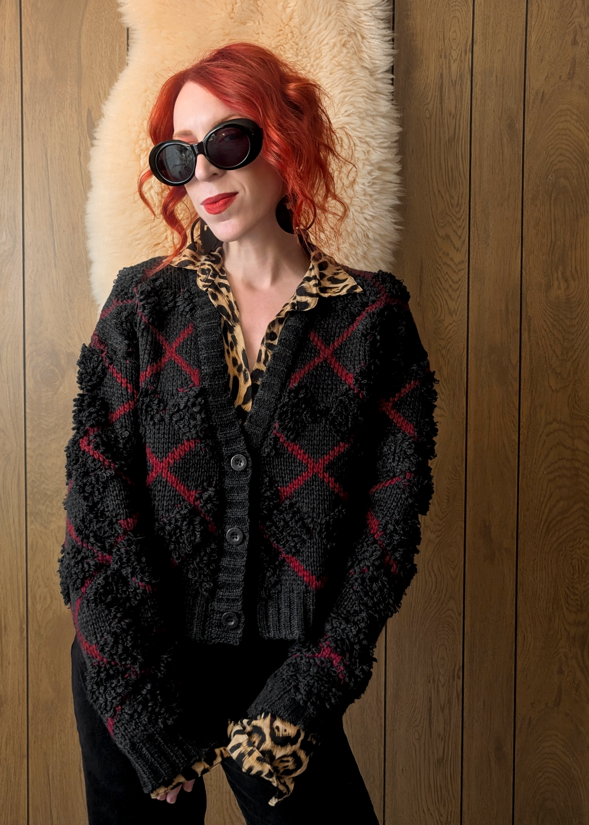 Person wearing a charcoal and red argyle "x" patterned cardigan over a leopard print shirt against a wooden wall.